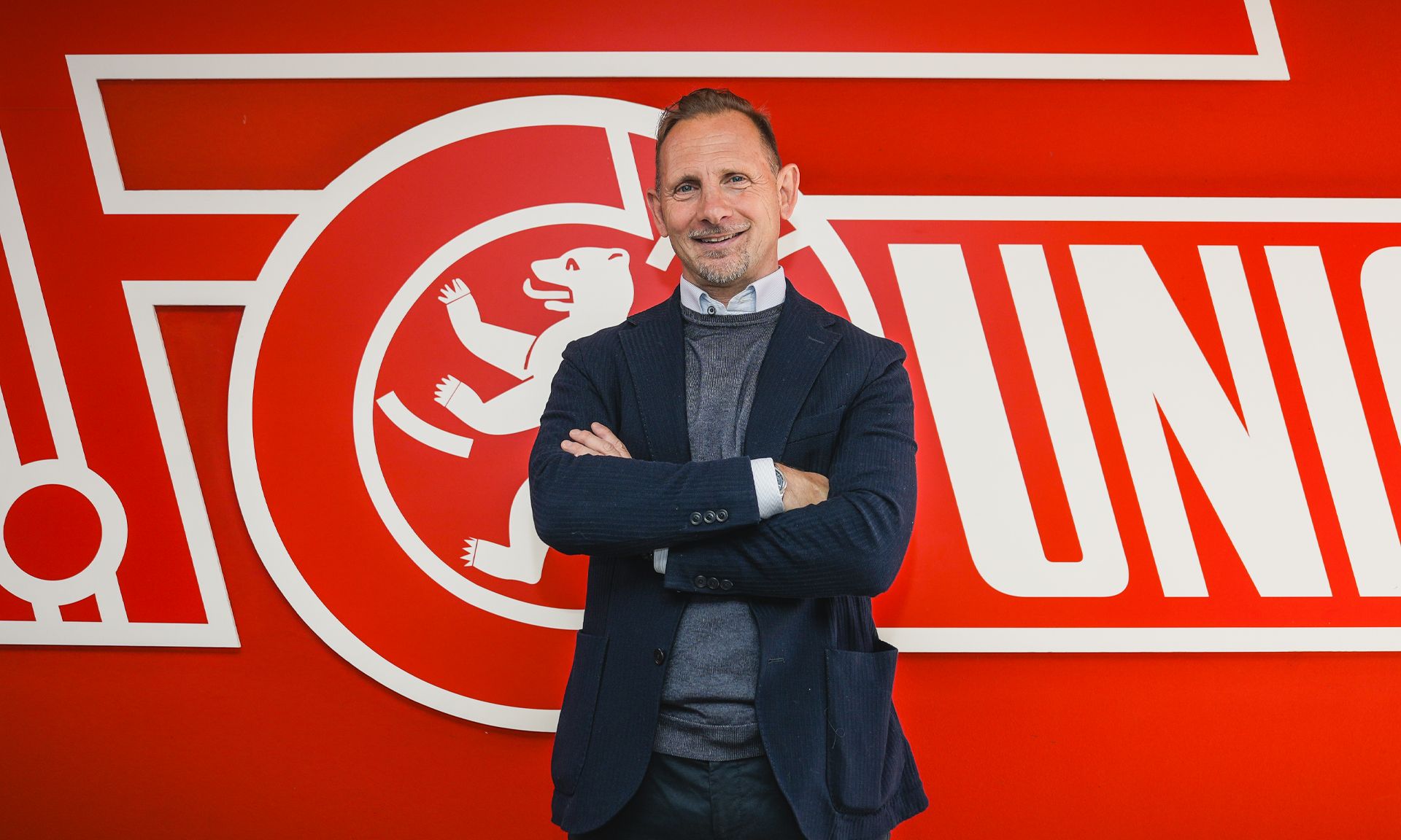 Mann in Anzug steht vor rotem Hintergrund mit dem Logo eines Vereins, lächelt und hat die Arme verschränkt.