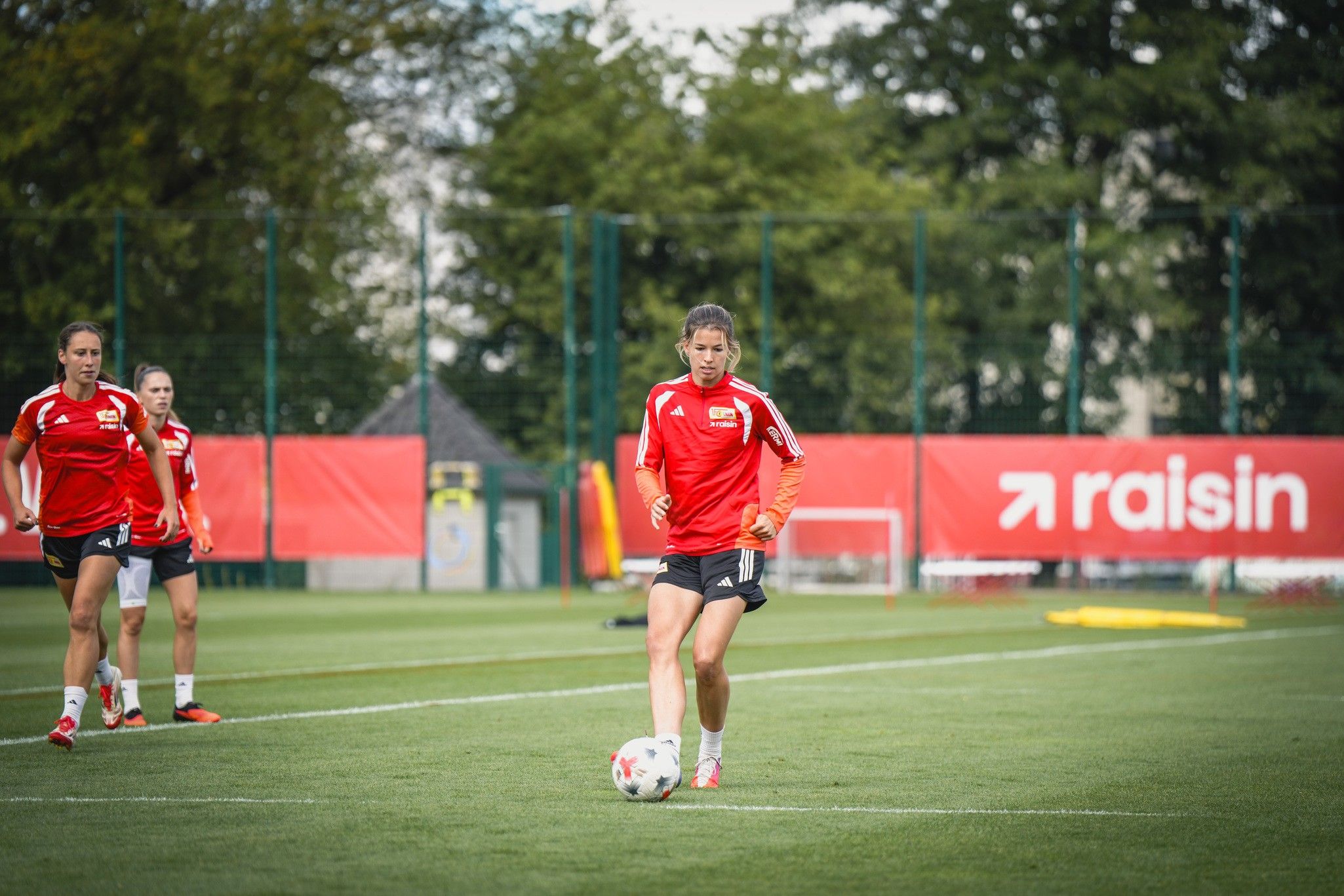 Eine Fußballspielerin in rotem Trikot dribbelt den Ball auf einem Trainingsplatz, im Hintergrund weitere Spielerinnen.