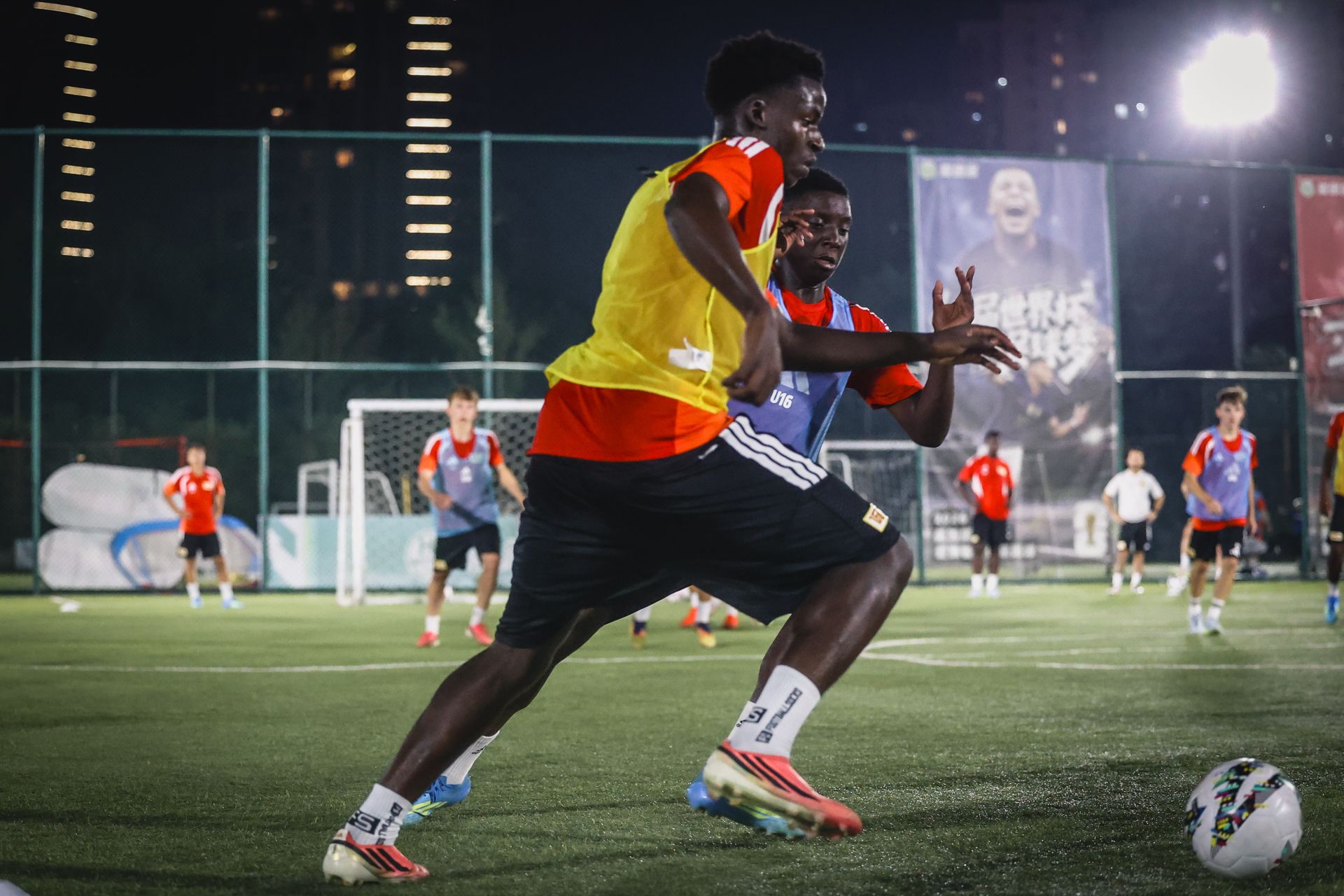 Zwei junge Fußballspieler in Training, einer im gelben, der andere im roten Shirt, dribbeln auf einem Platz mit anderen Spielern im Hintergrund.