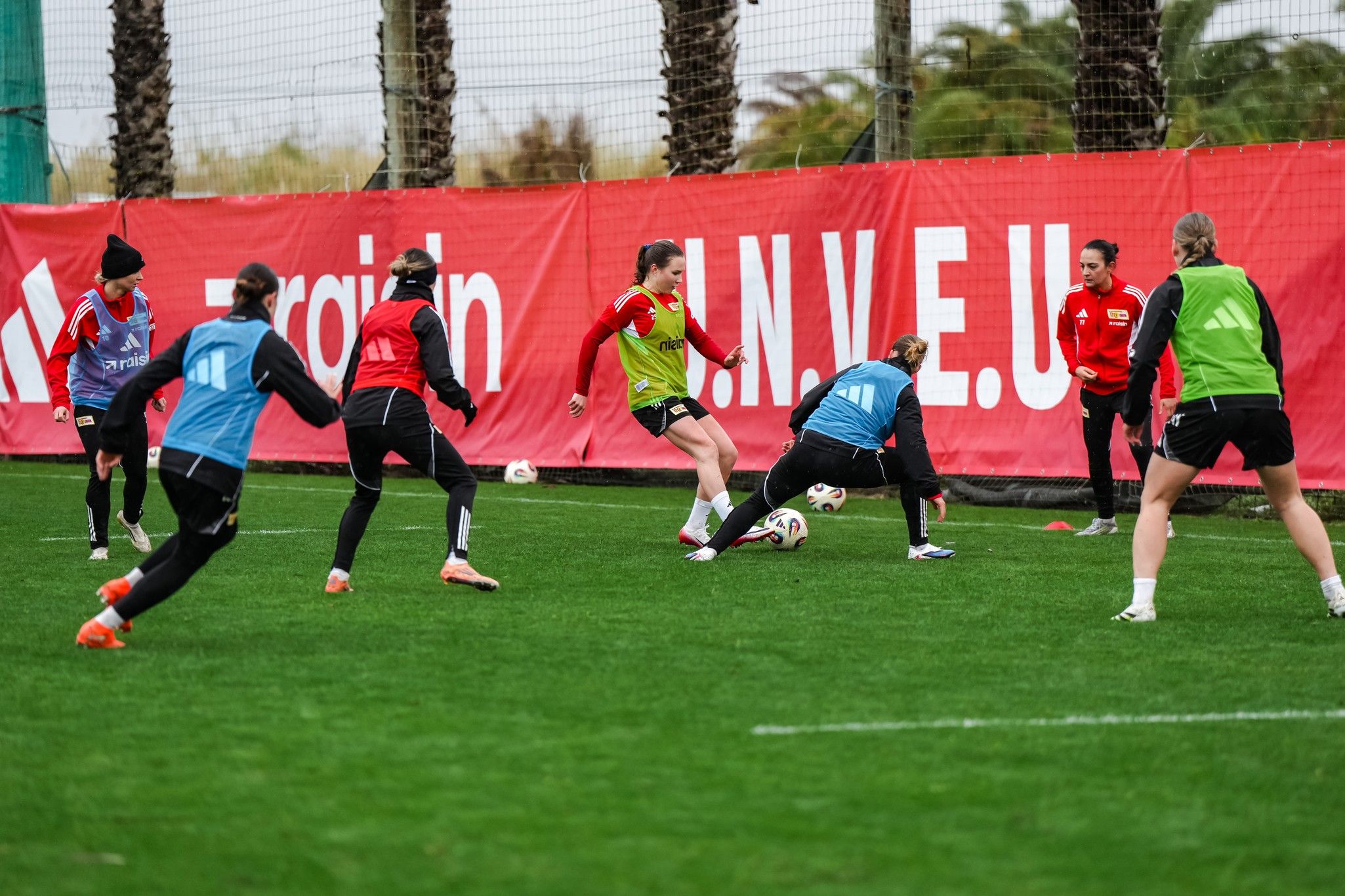 Frauenfußballmannschaft beim Training auf dem Kunstrasen, mehrere Spielerinnen in Aktion, rote Banner im Hintergrund.