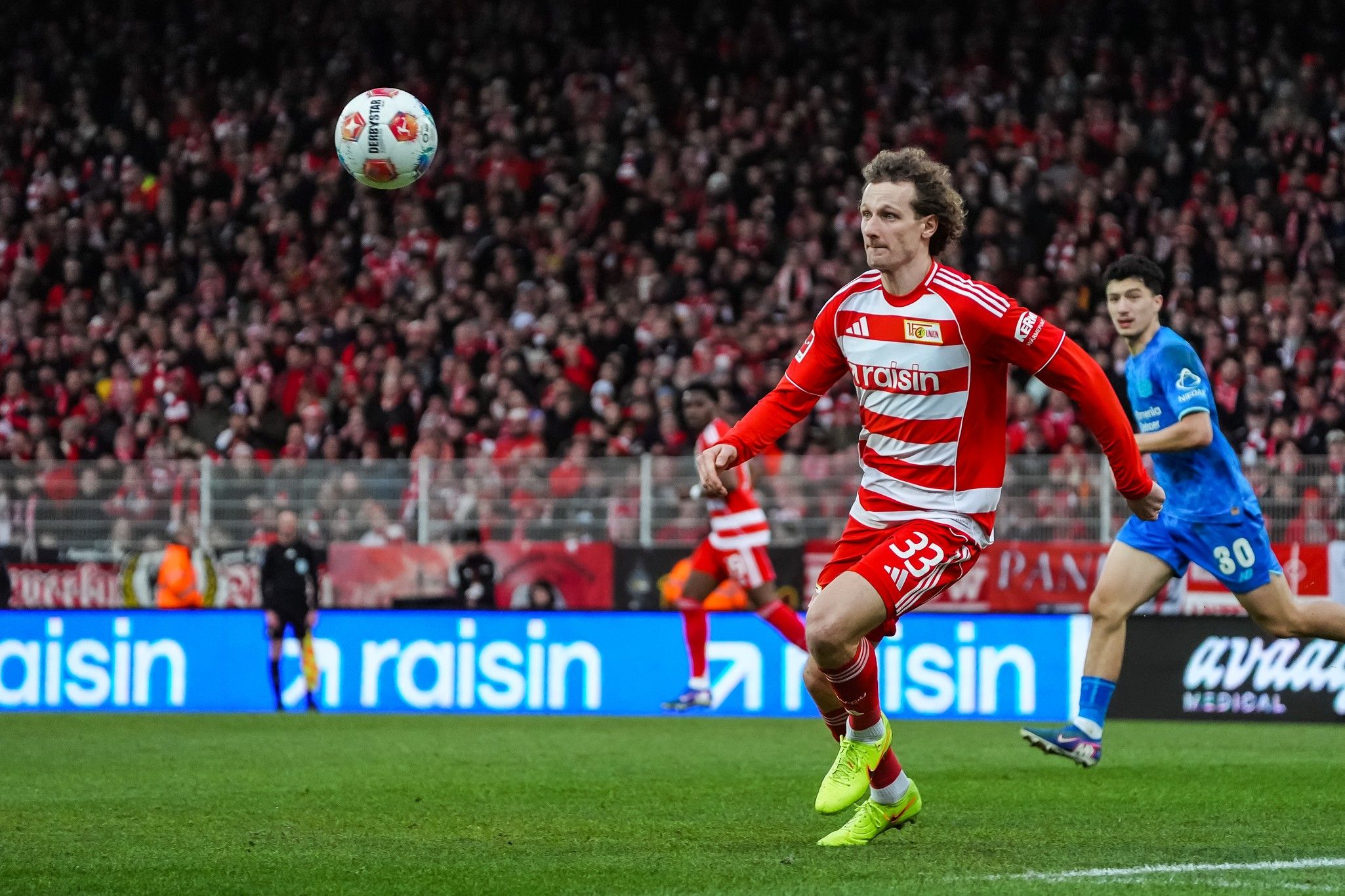 Ein Spieler in rot-gestreiftem Trikot sprintet über das Spielfeld, während ein Ball in der Luft fliegt und Zuschauer im Hintergrund jubeln.