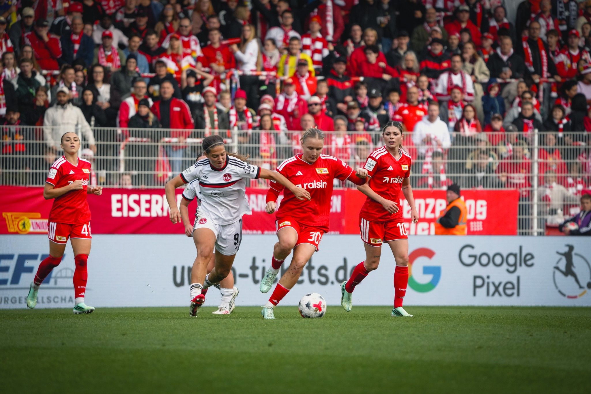 Eine Spielerin im weißen Trikot dribbelt den Ball, während zwei Spielerinnen in roten Trikots sie herausfordern. Stadion mit Fans im Hintergrund.