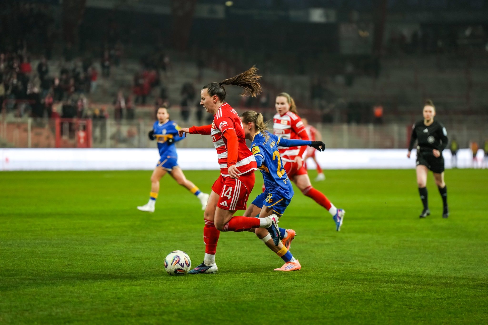Zwei Fußballspielerinnen in roten und blauen Trikots kämpfen um den Ball auf einem Rasenplatz während eines Spiels.