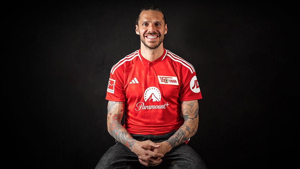 Smiling man in a red football jersey with club logo, sitting in front of a black background.