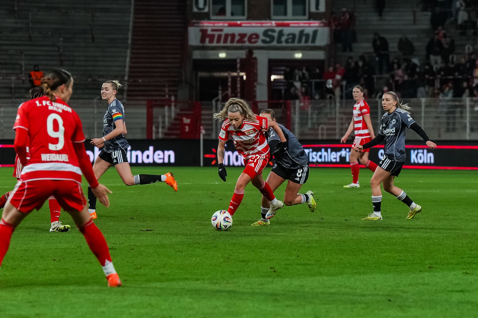 Spielszene eines Fußballspiels mit mehreren Spielerinnen, die um den Ball kämpfen, während Zuschauer im Hintergrund zu sehen sind.