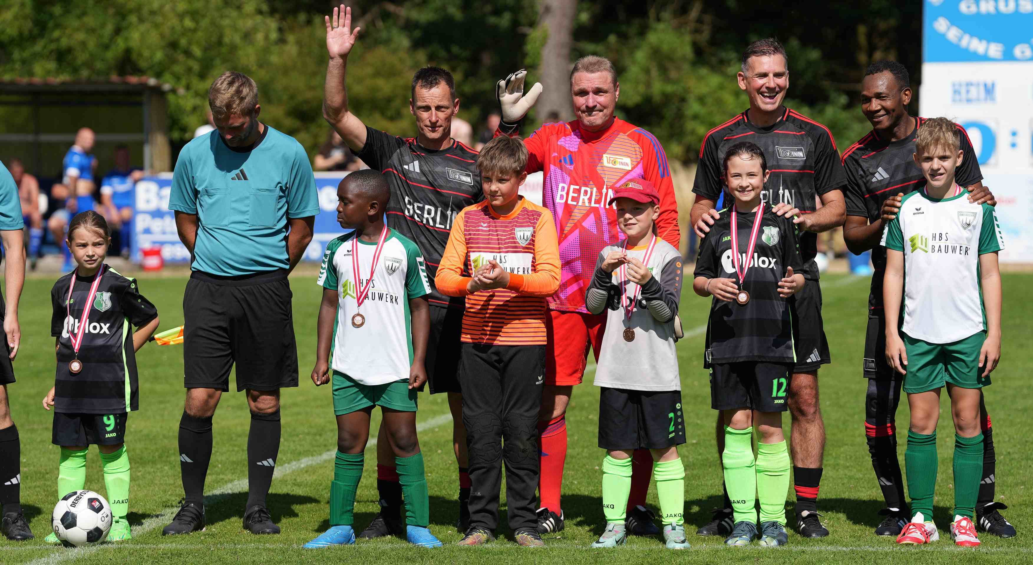 Fußballmannschaften stehen auf einem Platz, einige Spieler tragen Medaillen. Ein Torwart winkt, während andere lächeln.