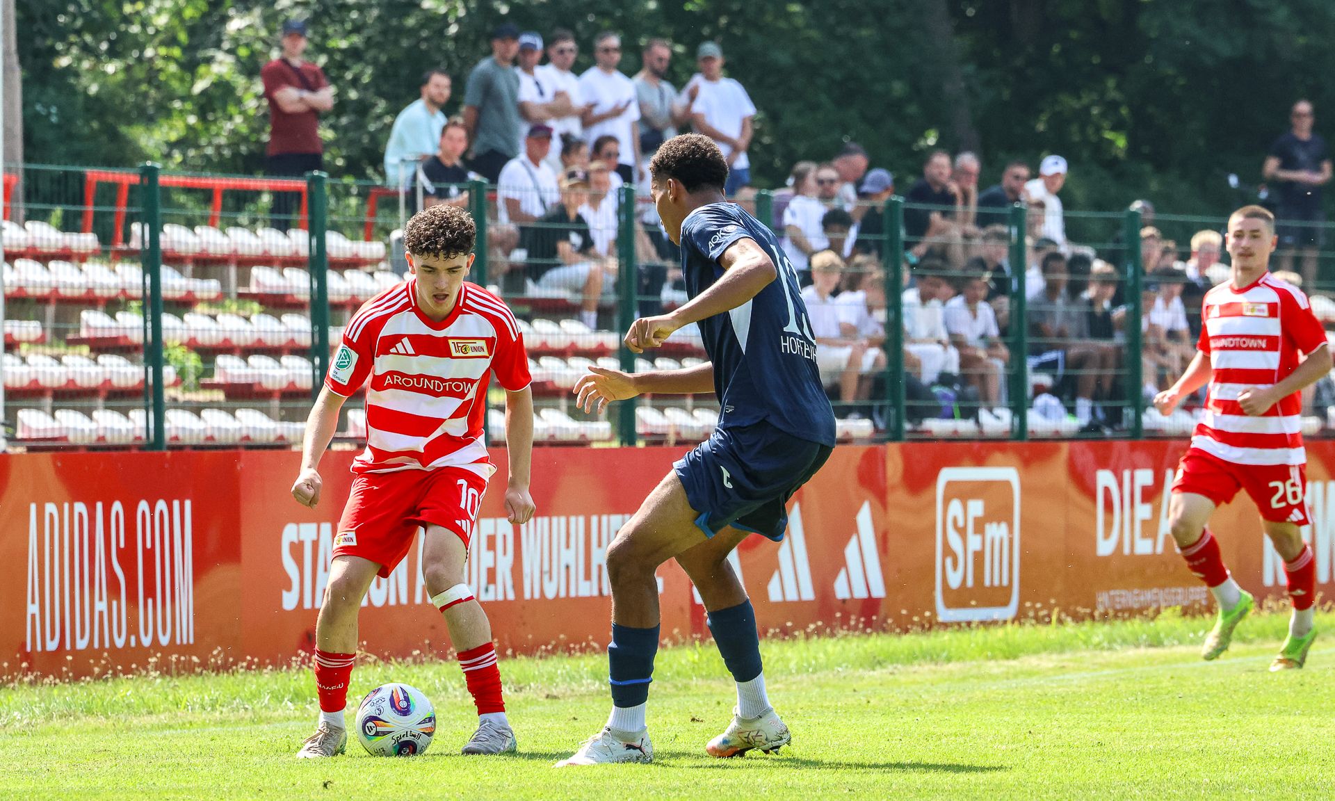 Ein Fußballspieler in rotem Trikot dribbelt den Ball, während ein Gegner in dunkelblauem Trikot ihn angreift. Zuschauer im Hintergrund.
