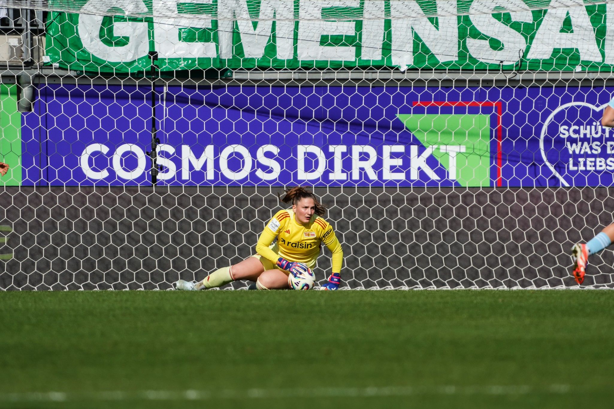 Eine Torhüterin sitzt hinter dem Tor mit einem Netz im Hintergrund, während ein Spieler in blauer Ausrüstung sich nähert.
