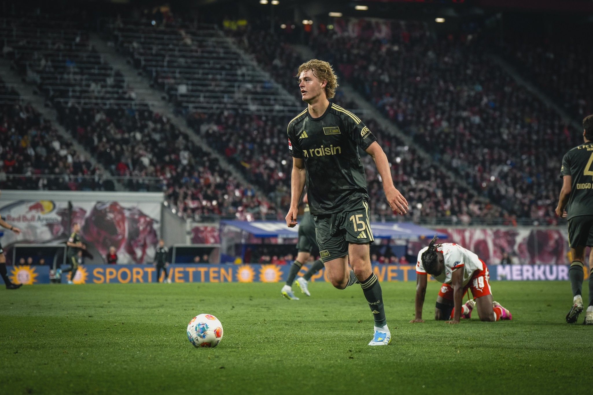 Ein Fußballspieler in grauem Trikot läuft über das Feld, während ein anderer Spieler hinter ihm am Boden liegt.