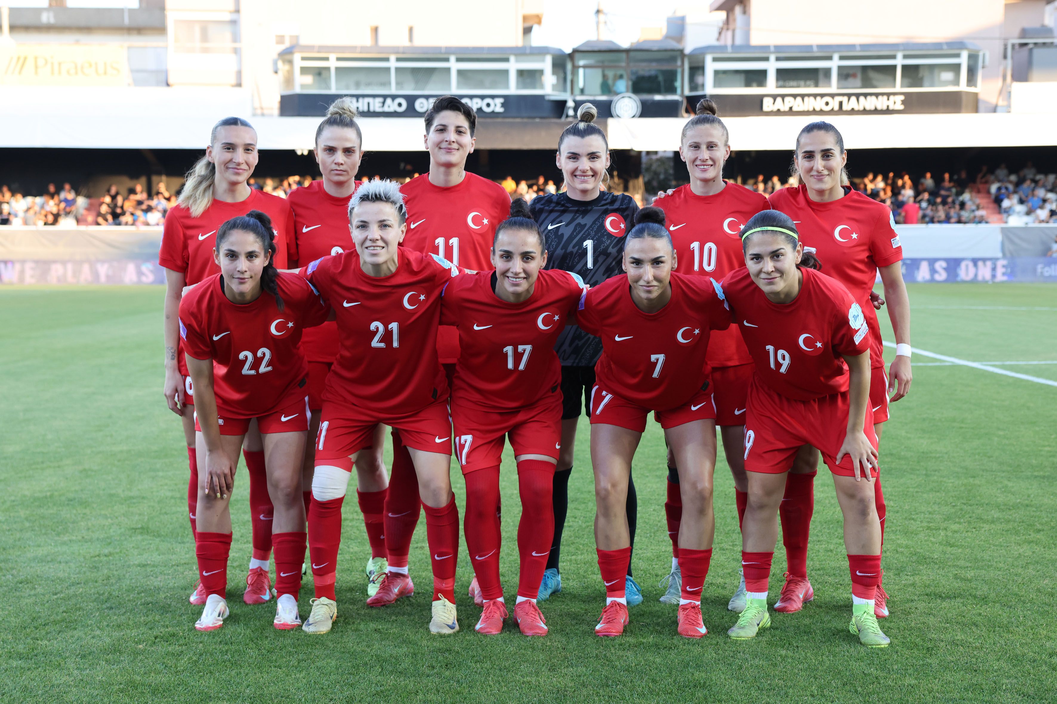 Eine Frauenfußballmannschaft in roten Trikots mit türkischem Wappen, auf einem Sportplatz vor einer Zuschauertribüne.