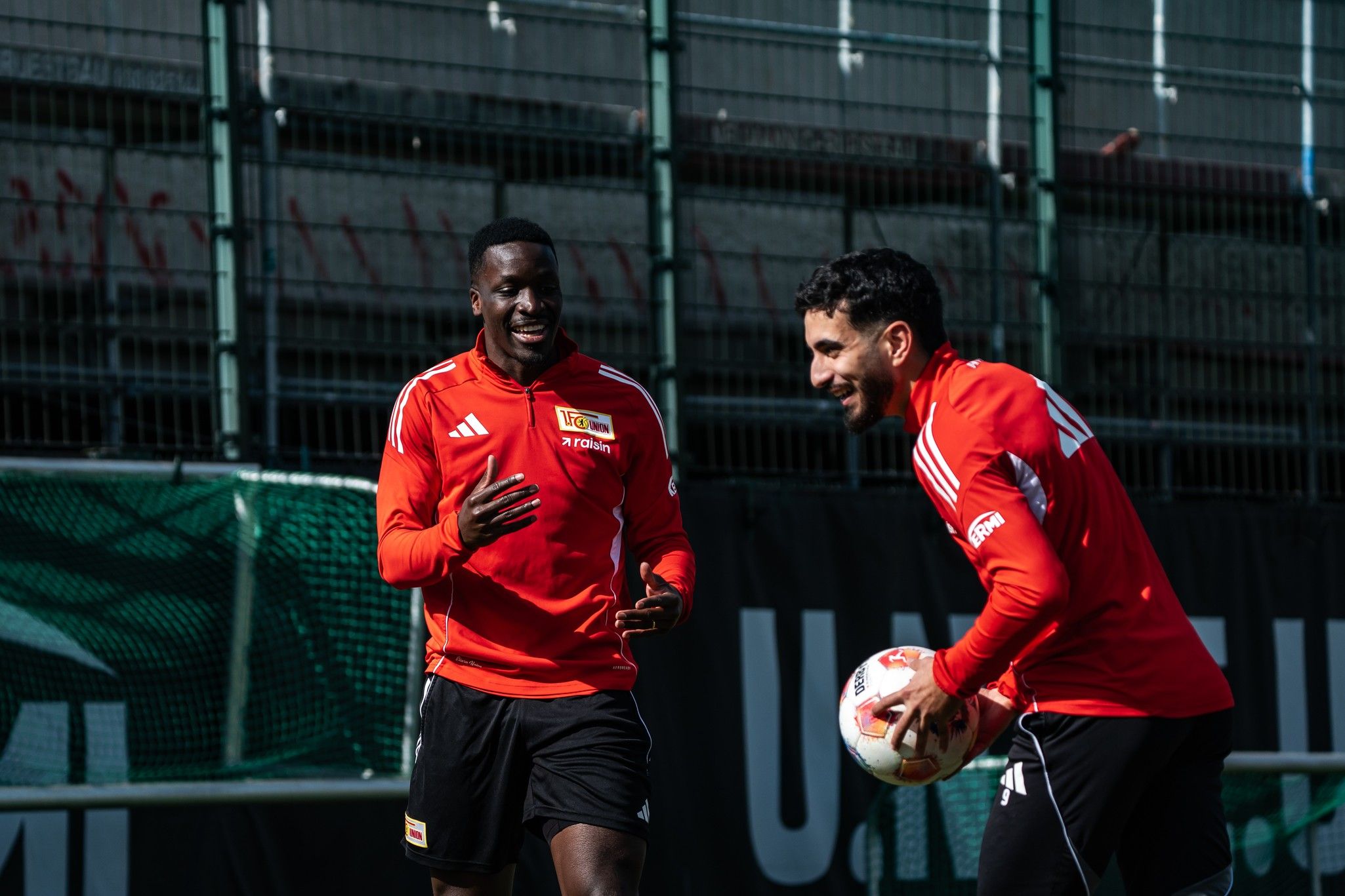 Zwei Fußballspieler in roten Trikots lachen und haben Spaß beim Training auf dem Platz.