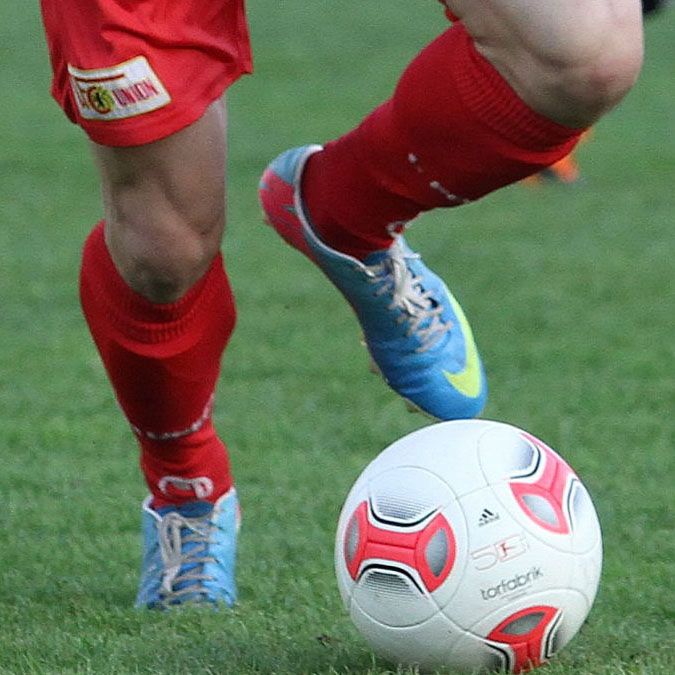 Ein Fußballspieler in roter Sportkleidung tritt einen weißen Fußball auf einem grünen Rasenfeld.