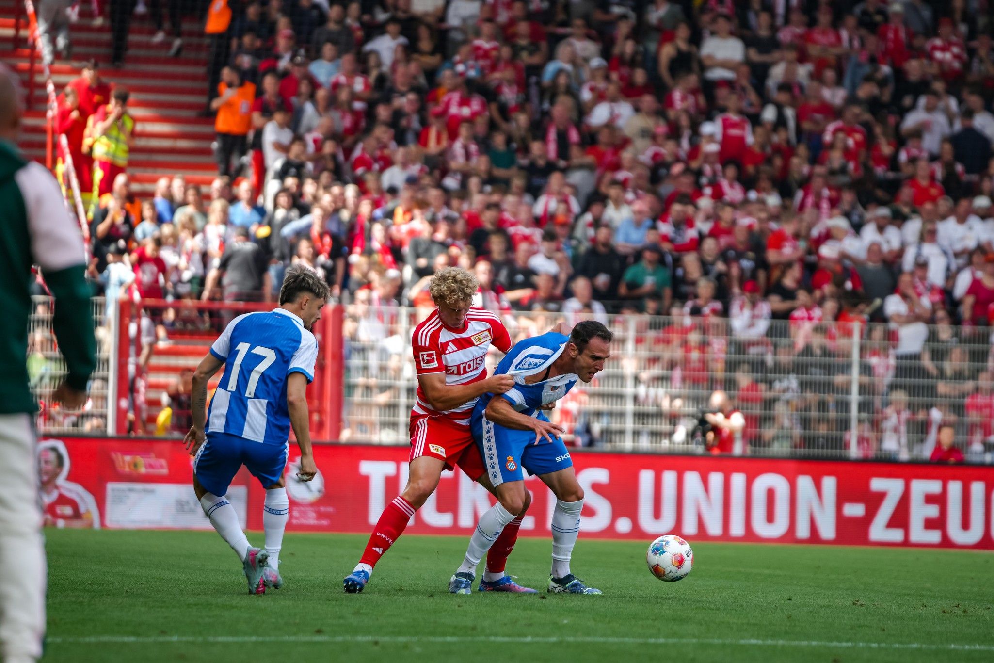 Ein Fußballspiel mit Spielern in blauen und roten Trikots, die um den Ball kämpfen, vor einer jubelnden Menschenmenge.