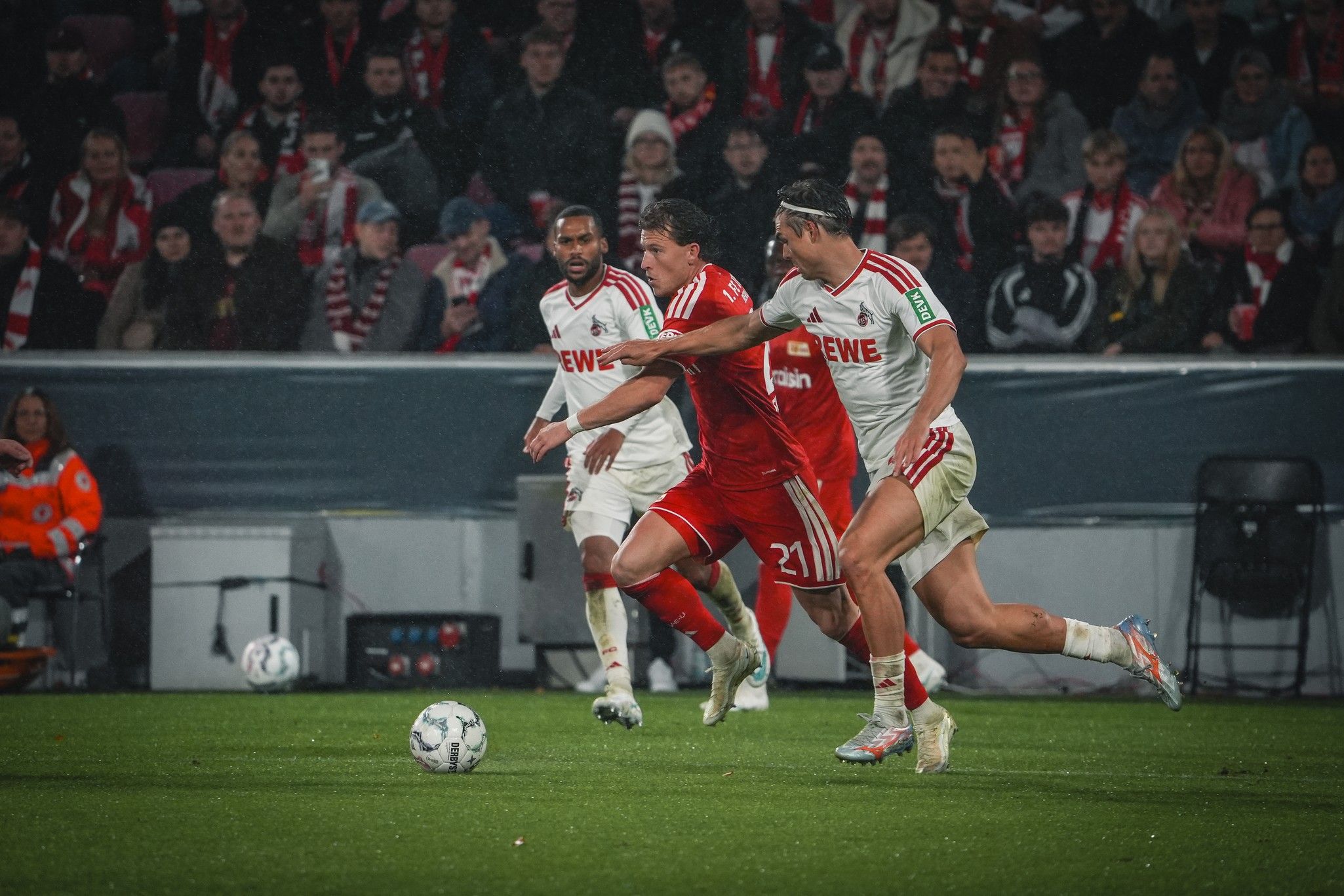 Zwei Fußballspieler in roten und weißen Trikots kämpfen um den Ball auf einem nassen Spielfeld, umgeben von Zuschauern.