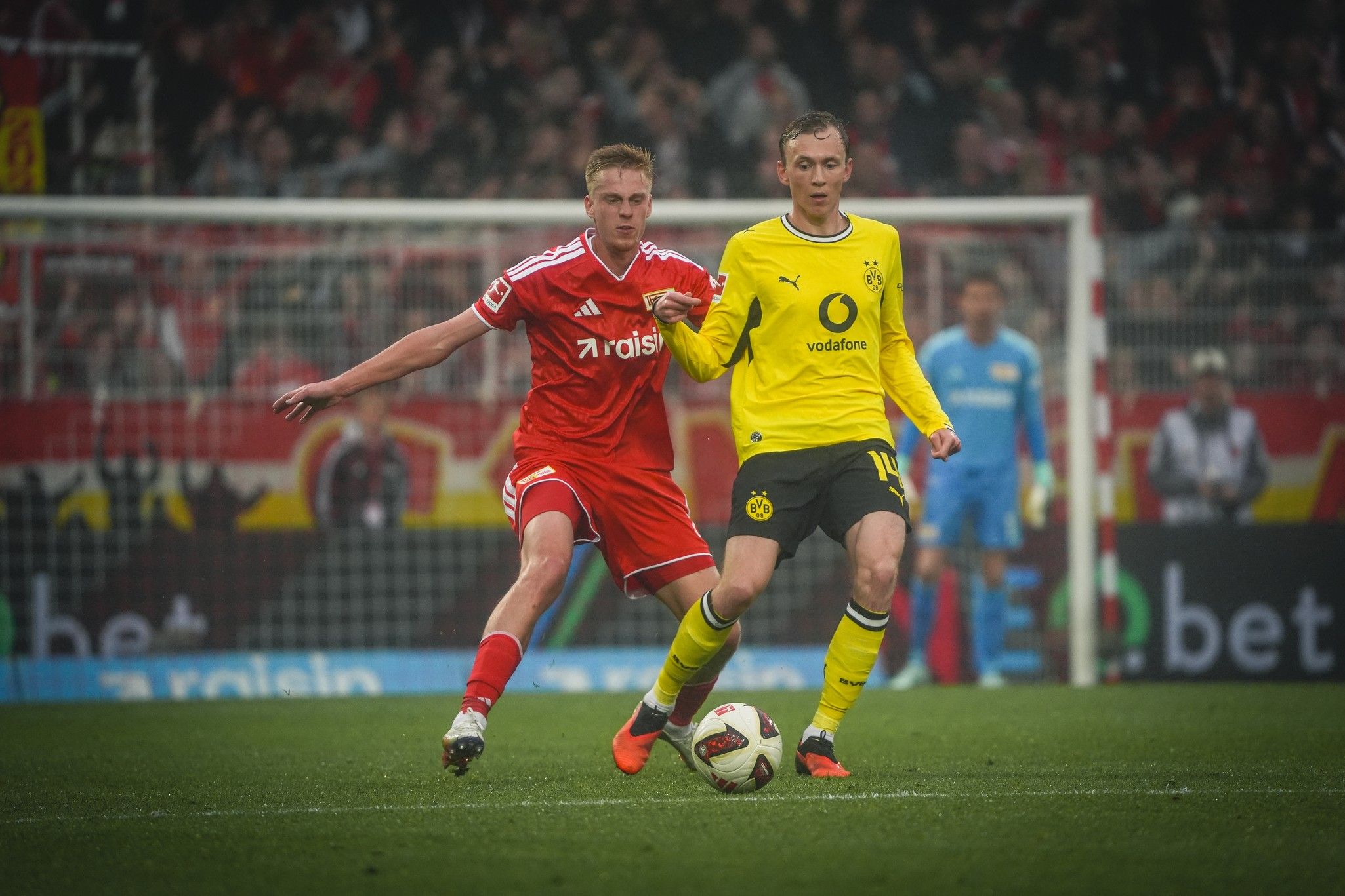 Fußballspiel zwischen zwei Spielern, einer in rotem und einer in gelbem Trikot, auf dem Spielfeld während eines Spiels.