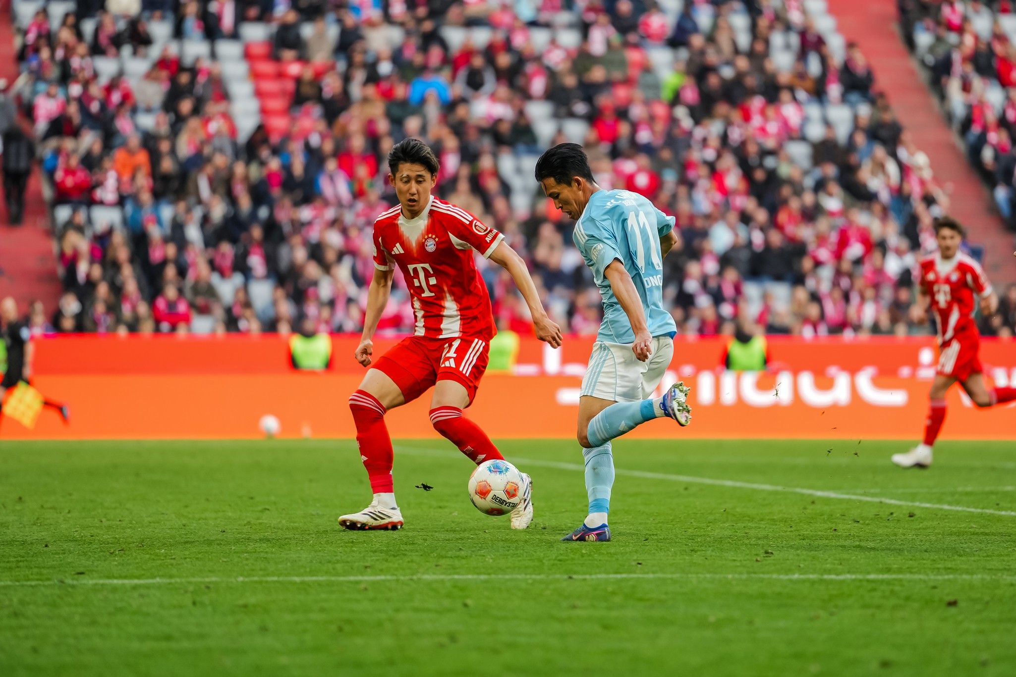 Spielszene zwischen zwei Fußballspielern, einer in rotem, der andere in hellblauem Trikot, im Stadion mit Zuschauern im Hintergrund.