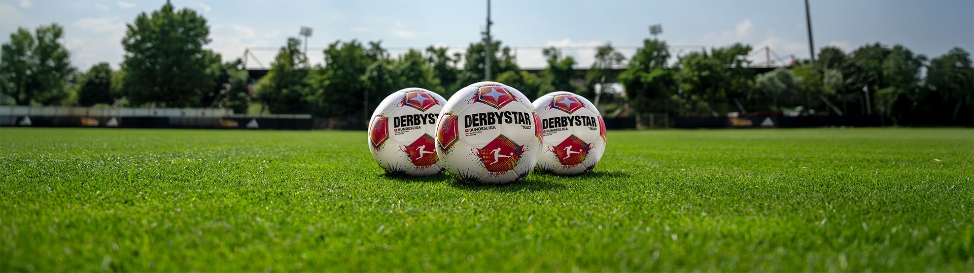 Drei Fußballbälle liegen auf einem frisch gemähten grünen Rasenfeld. Hintergrund mit Bäumen und einem Stadion.
