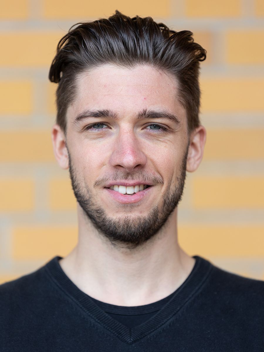 Junger Mann mit kurz geschnittenem Haar und Bart, lächelnd vor einer gelben Backsteinmauer.