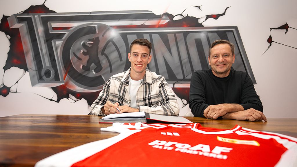 Ein junger Mann und ein älterer Mann sitzen an einem Tisch, der mit einem Sporttrikot und einen Vertrag bedeckt ist. Im Hintergrund ist ein Logo zu sehen.