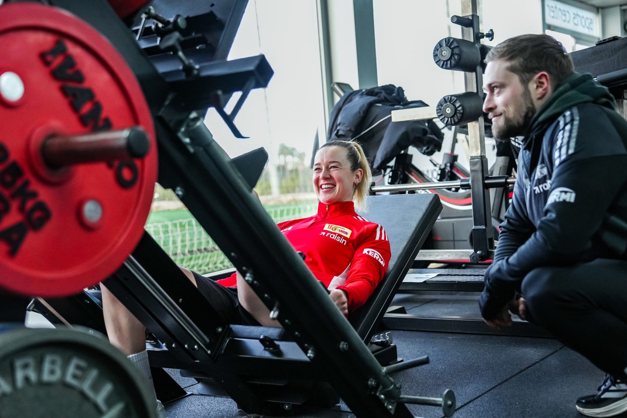 Eine Frau trainiert an einem Beinpresse-Gerät, während ein Trainer sie beobachtet und anfeuert.