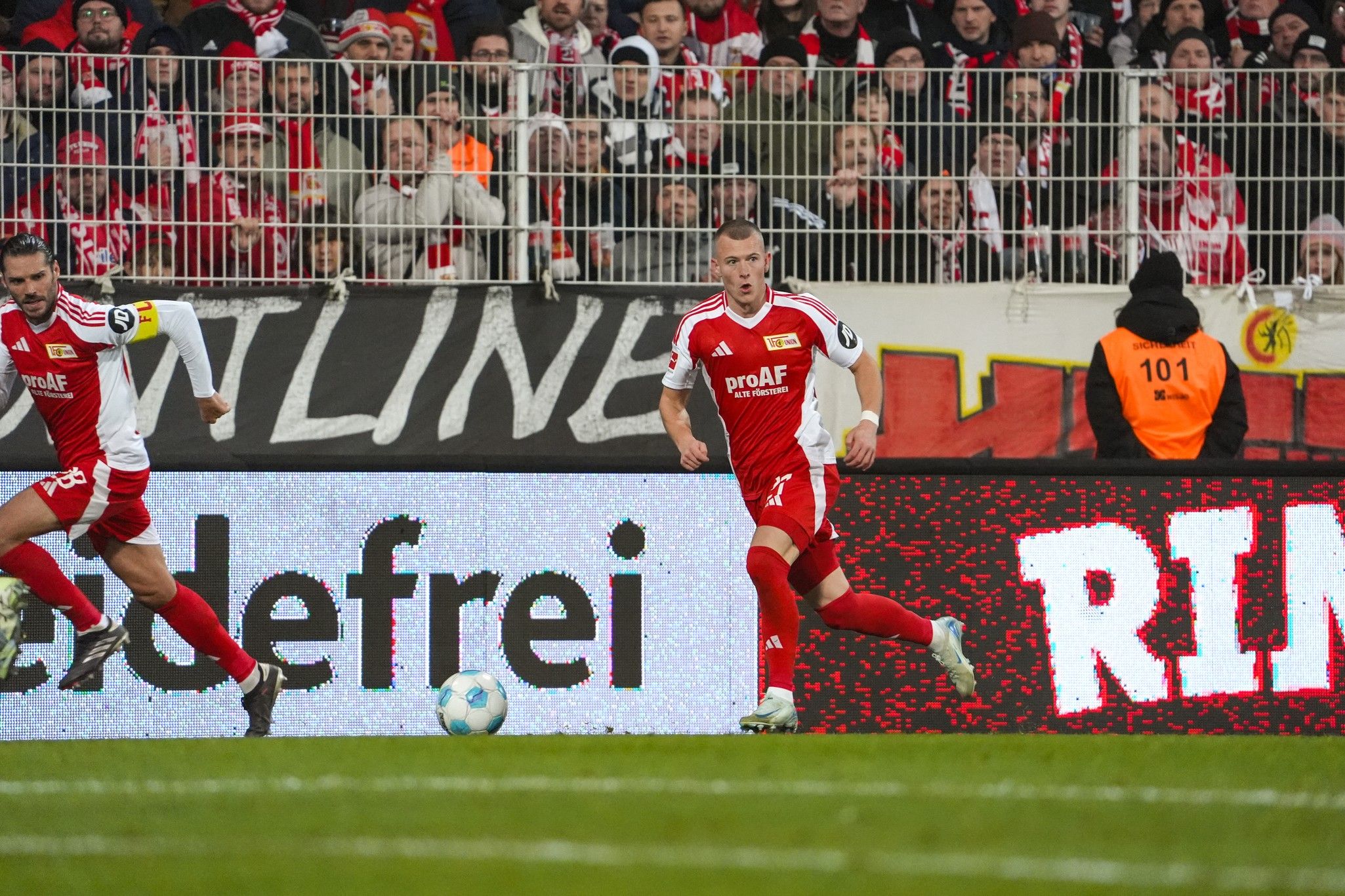 Game scene with two soccer players in red jerseys facing an opponent, with a cheering crowd in the background.