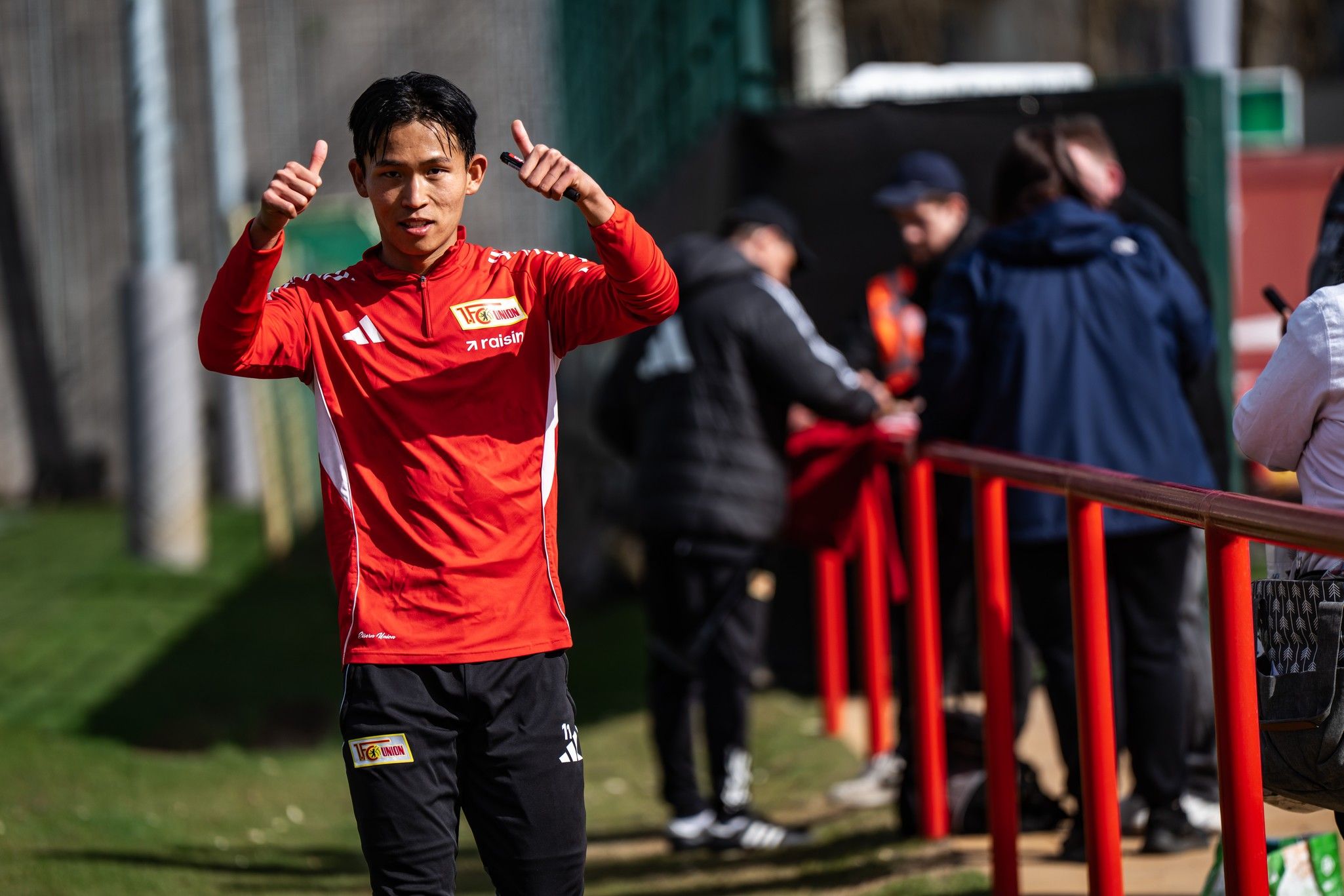 Ein Sportler in rotem Trikot zeigt mit beiden Daumen nach oben und lächelt. Im Hintergrund sind weitere Personen und ein Fußballplatz zu sehen.