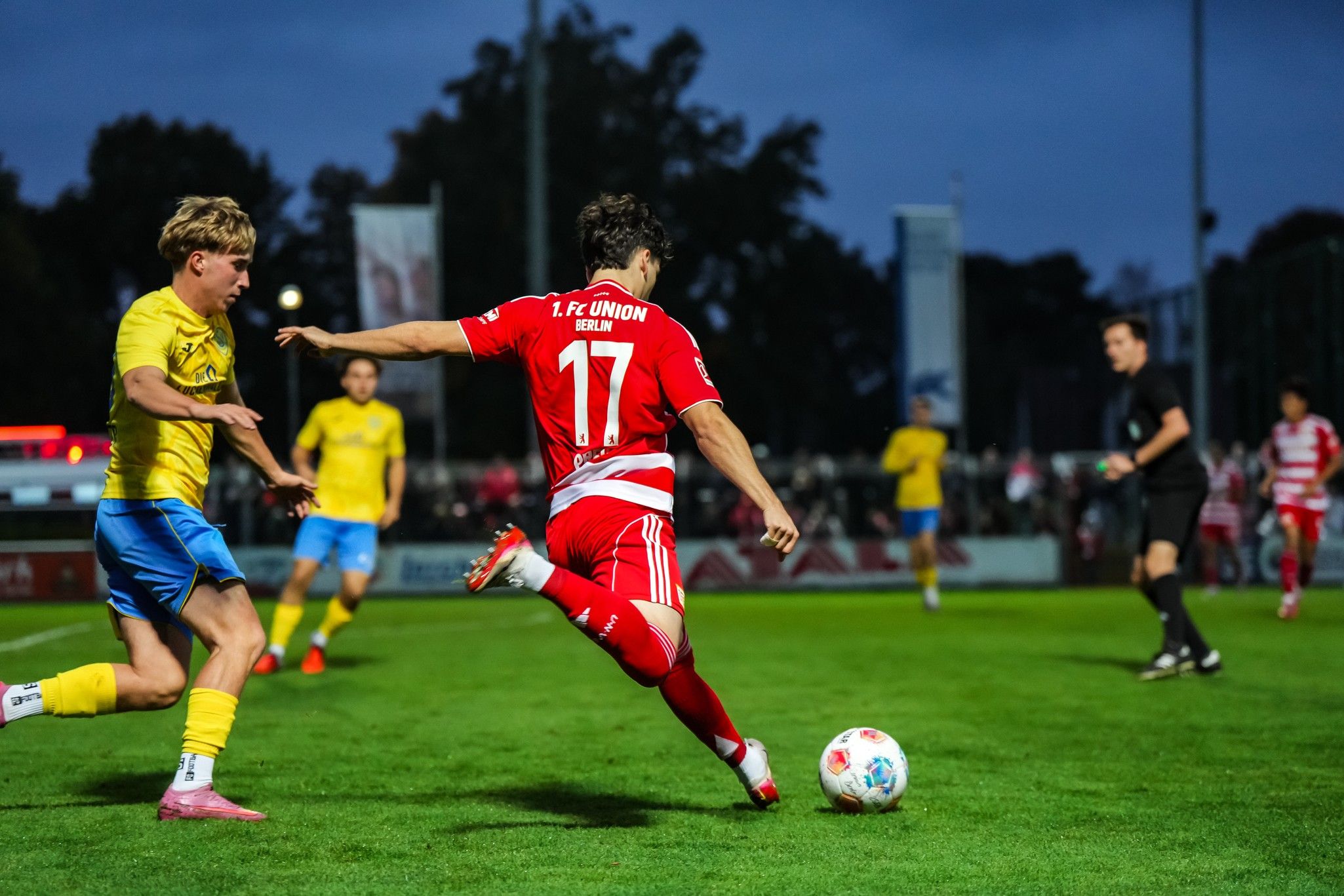 Spielszene im Fußball: Ein Spieler in rotem Trikot tritt den Ball, während ein Gegner in gelbem Trikot ihn verfolgt.
