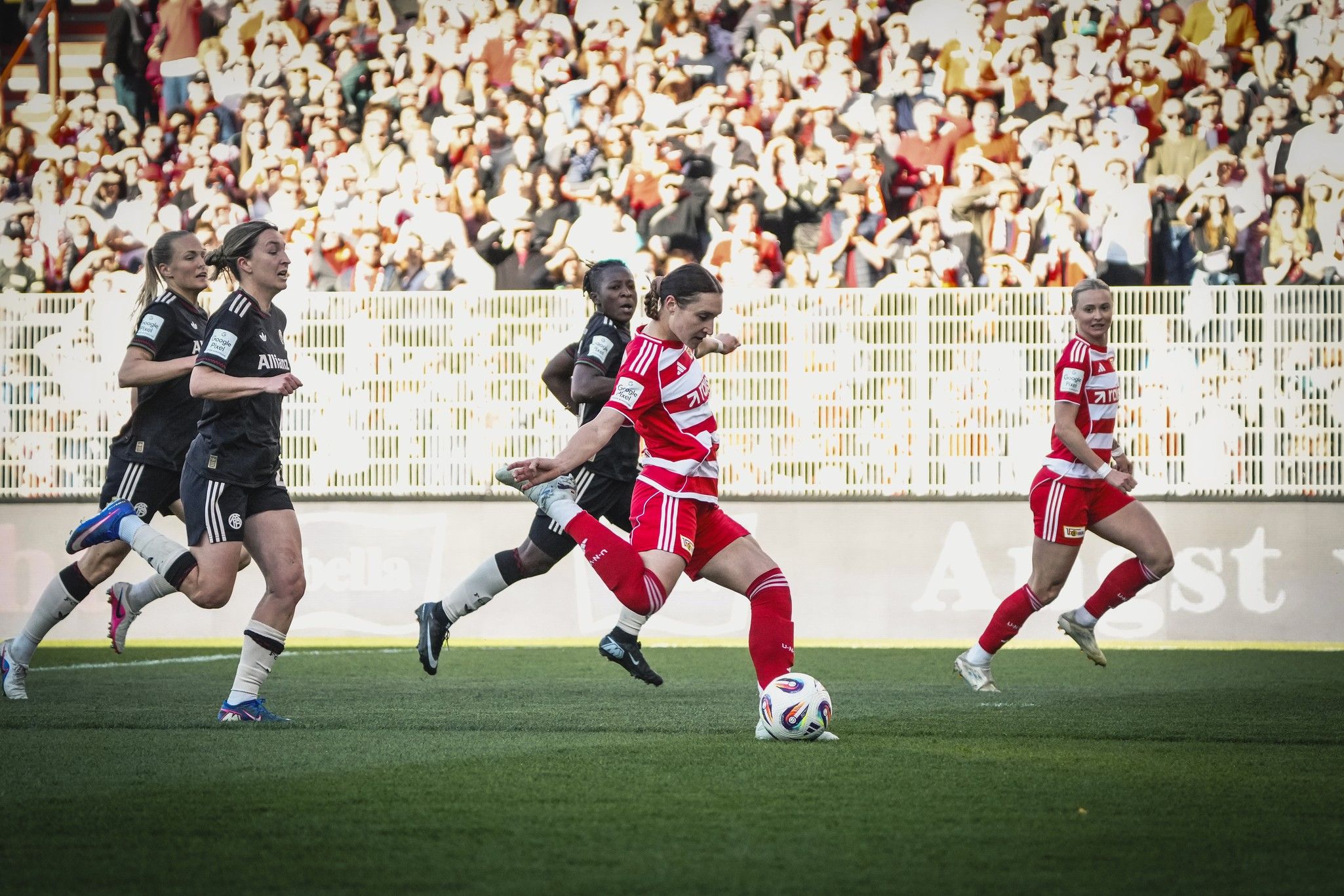 Eine Spielerin in rotem Trikot dribbelt den Ball, während sie von mehreren Gegenspielerinnen verfolgt wird. Publikum im Hintergrund.