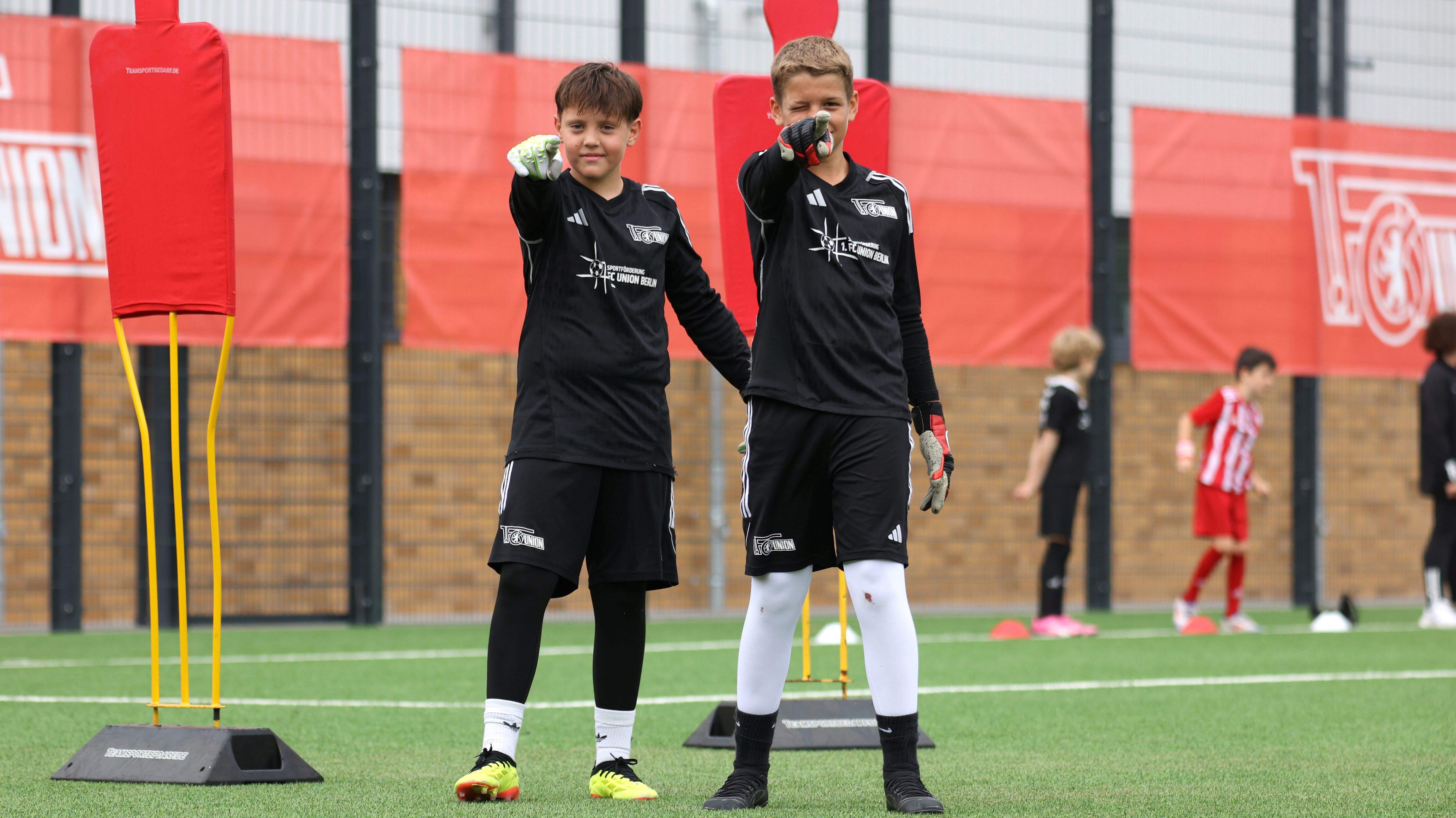 Zwei junge Fußballspieler in schwarzen Trikots stehen auf einem Trainingsplatz und zeigen mit dem Finger in die Kamera.