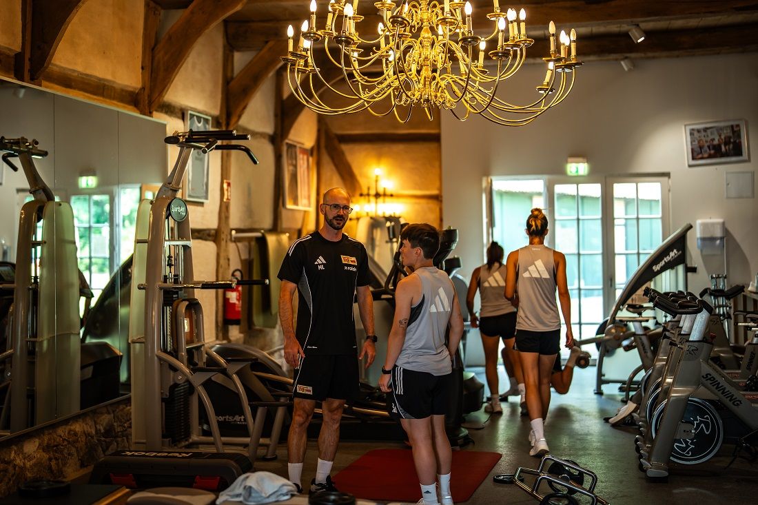 Personen in Sportkleidung unterhalten sich in einem Fitnessraum mit Gewichten und Trainingsgeräten, beleuchtet von einem Kronleuchter.
