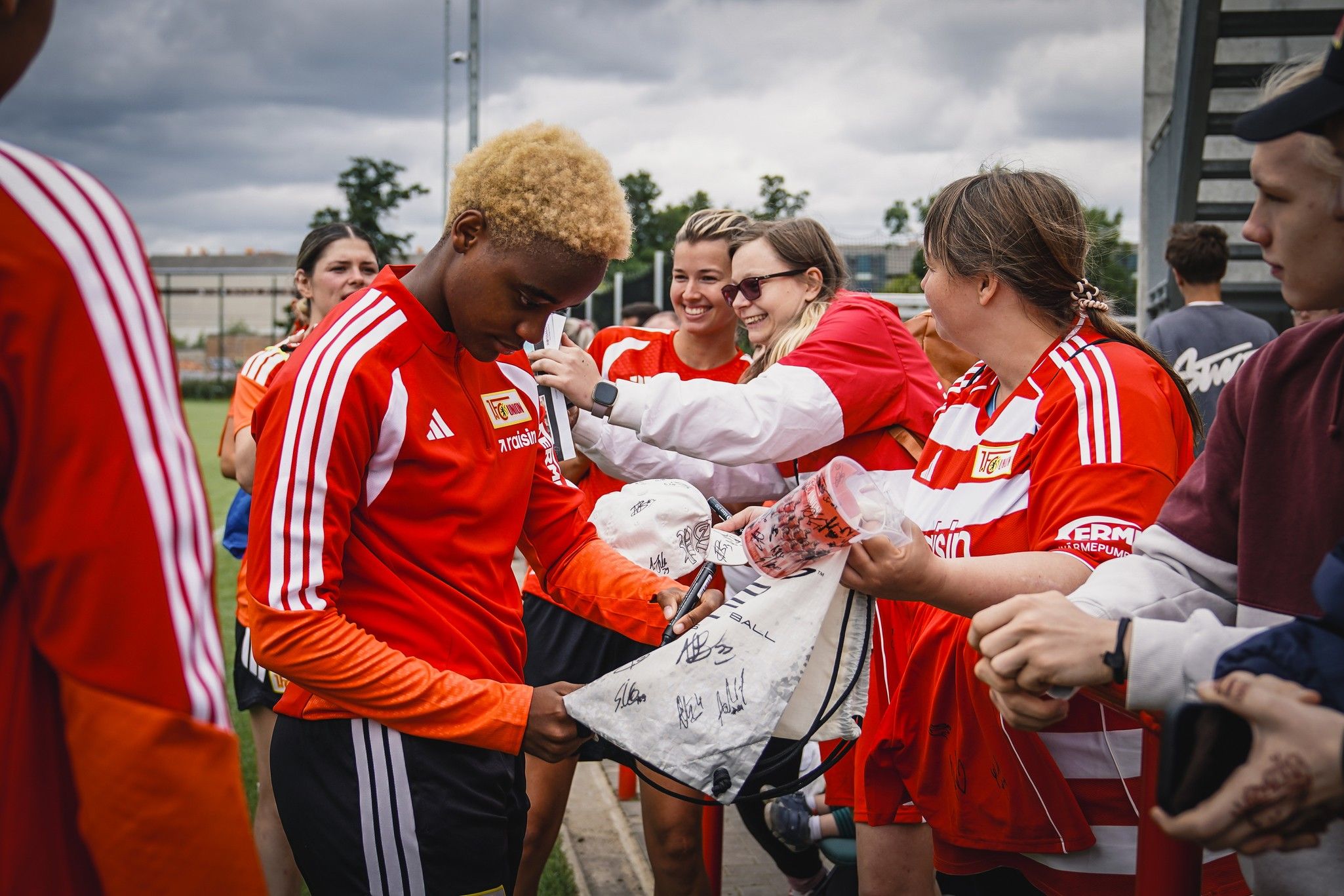 Eine Fußballspielerin mit blonden Locken signiert Autogramme für Fans in einem Stadion.