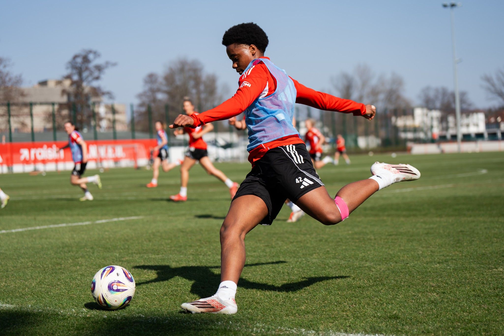Spieler trainiert auf dem Fu ßballplatz, während andere im Hintergrund üben. Sonne scheint über das Trainingsgelände.