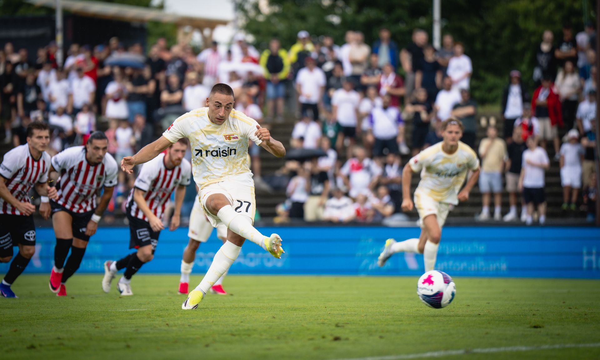 Fußballspieler schießt einen Elfmeter auf das Tor, im Hintergrund jubelnde Fans und Mitspieler.