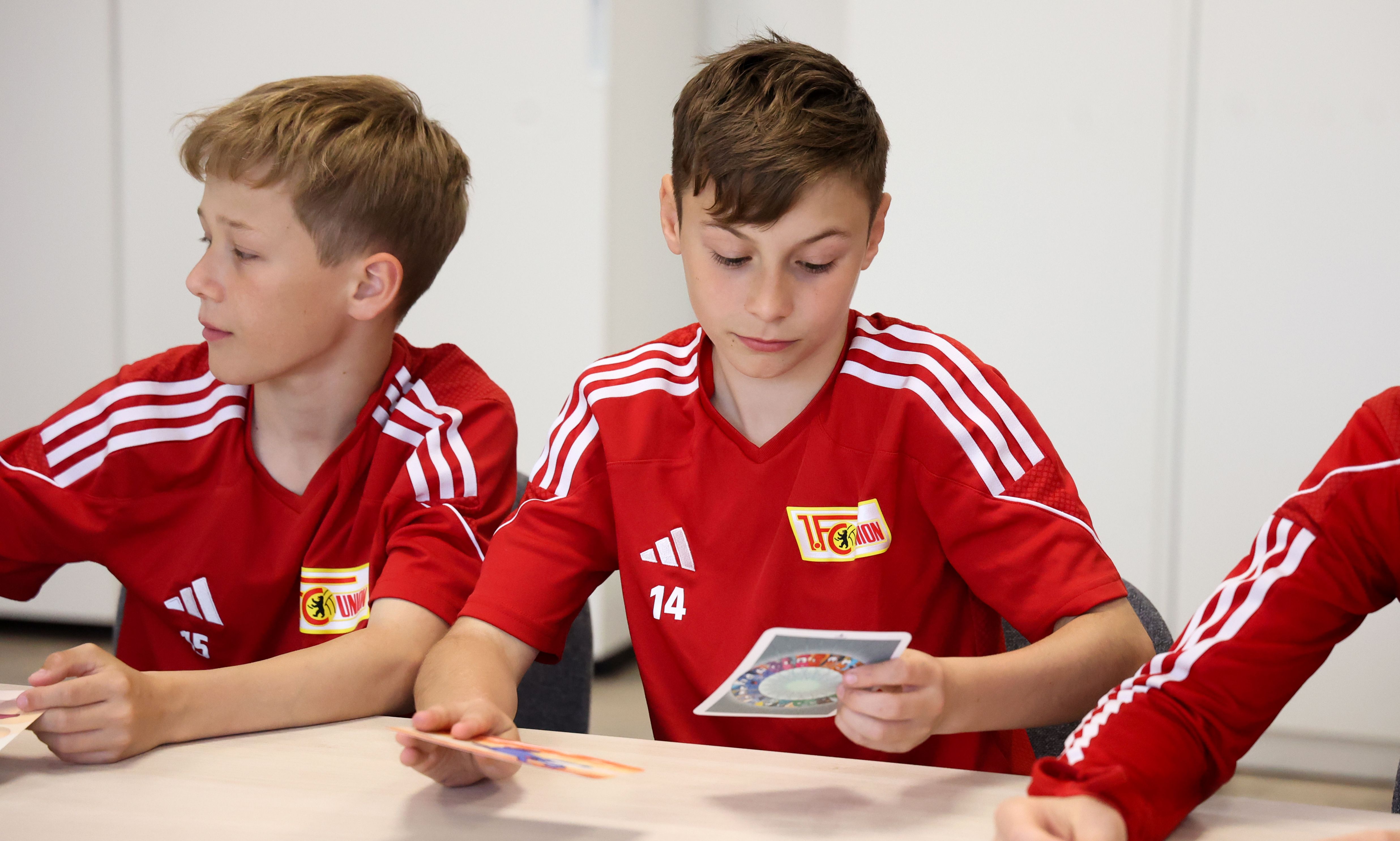 Drei Jungen in roten Trikots sitzen an einem Tisch und betrachten Karten.