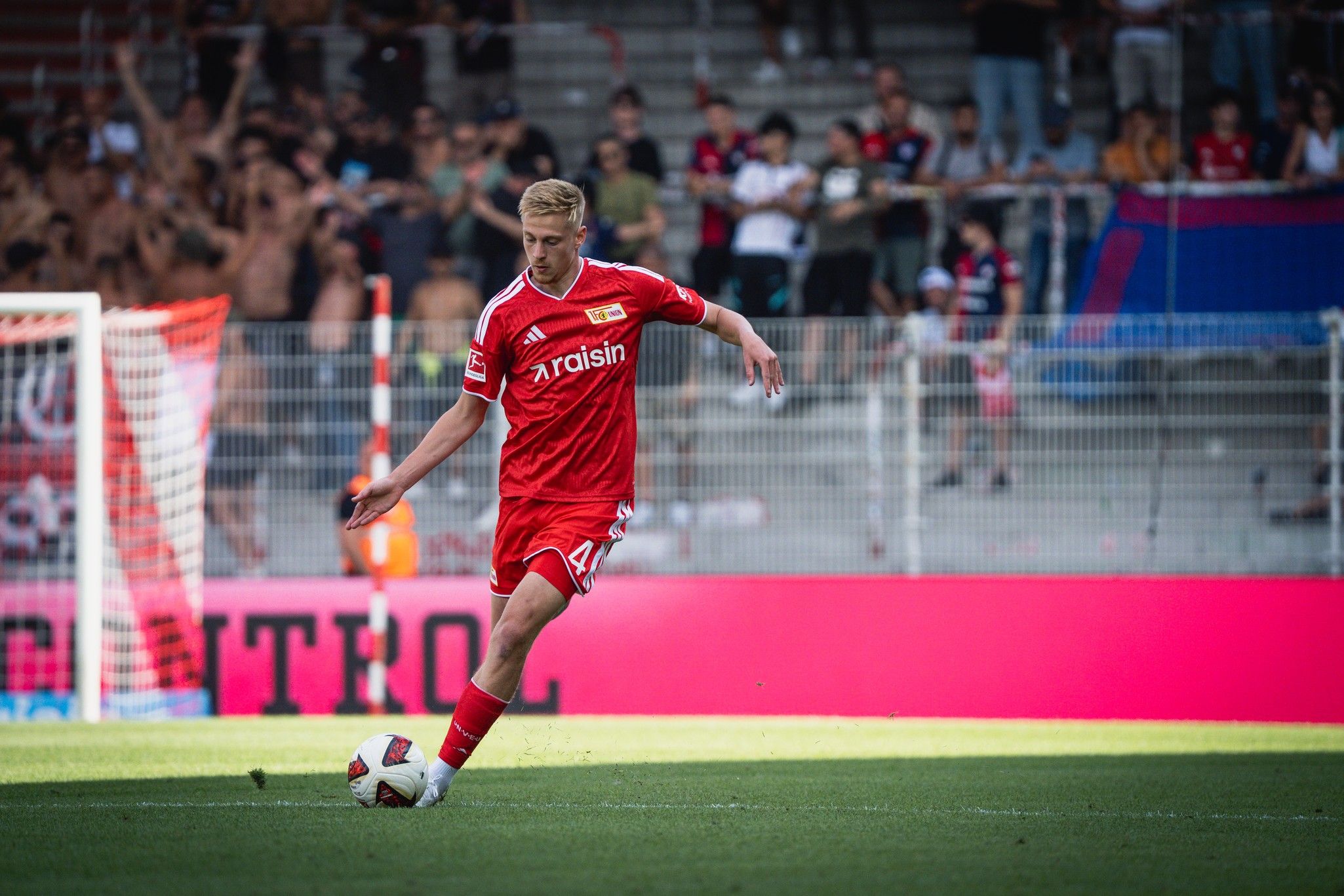 Ein Spieler in rotem Trikot führt einen Schuss auf das Tor aus, im Hintergrund jubelnde Fans in einem Stadion.