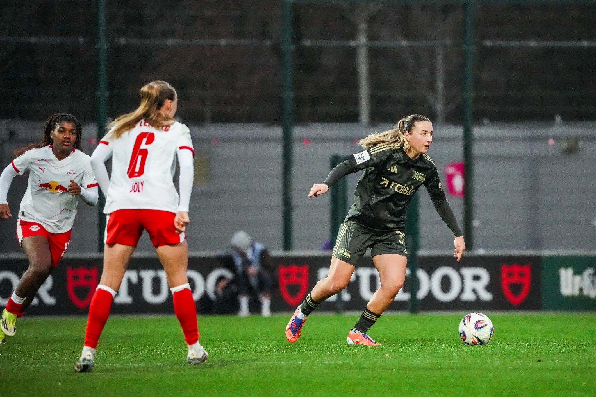 Eine Spielerin im schwarz-gelben Trikot dribbelt den Ball, während zwei Gegnerinnen in rot-weißen Trikots hinter ihr stehen.