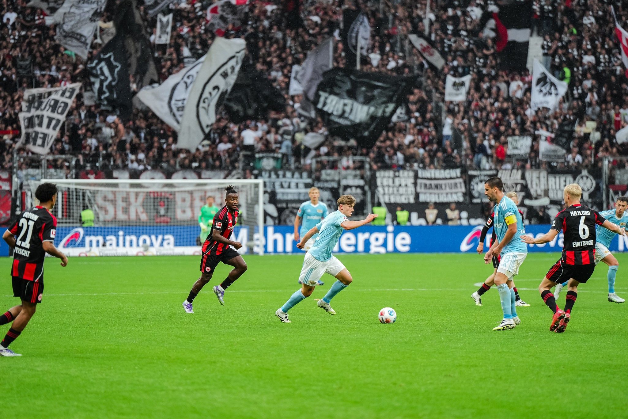 Zwei Fußballmannschaften spielen auf einem Rasenfeld, umgeben von jubelnden Fans mit Fahnen und Bannern im Hintergrund.