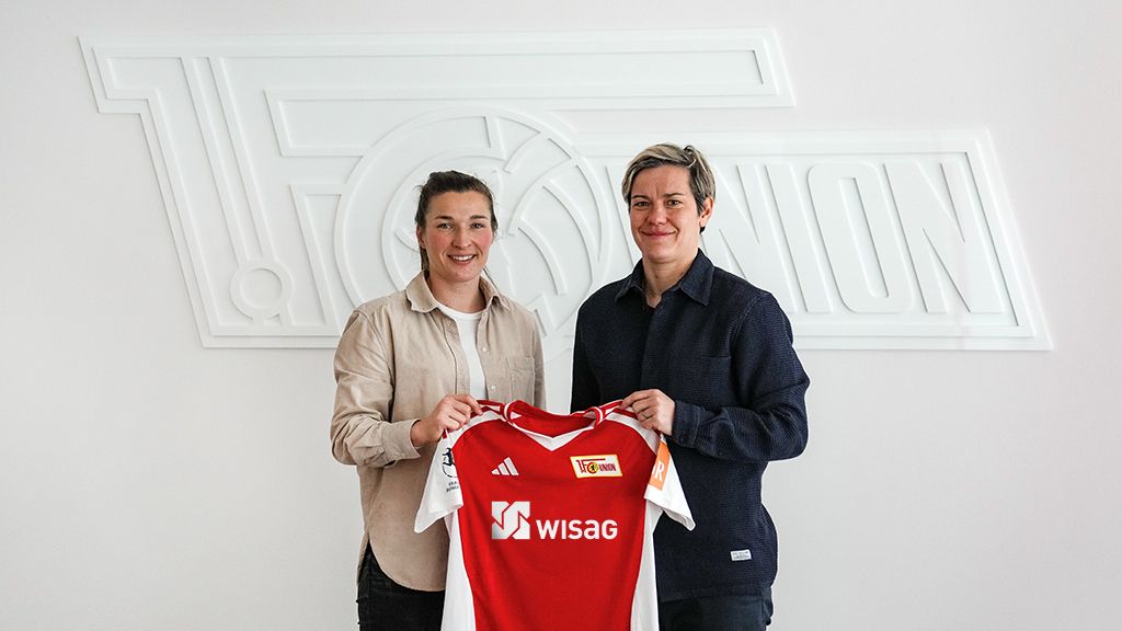 Zwei Personen halten ein Fußballtrikot mit dem Logo von 1. FC Union Berlin vor einer Wand mit dem Vereinsnamen.