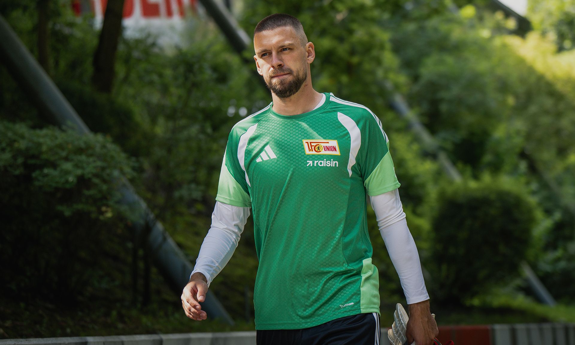 Fußballspieler in grünem Trainingsshirt und blauen Shorts, der auf einem Platz mit Bäumen im Hintergrund geht.
