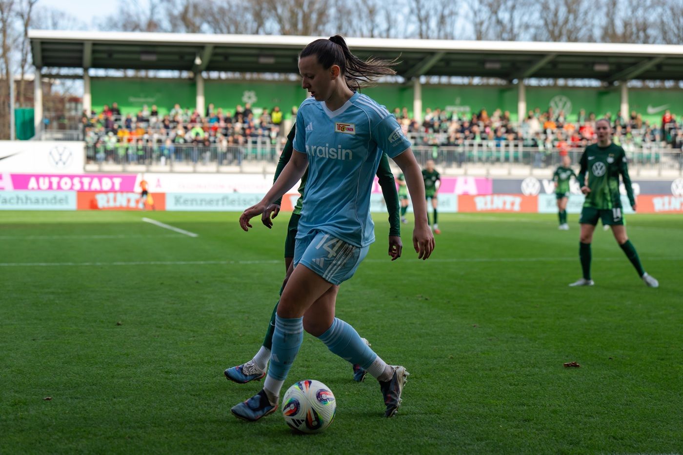 Eine Fußballspielerin in hellblauem Trikot dribbelt den Ball auf einem Rasenplatz mit Zuschauern im Hintergrund.