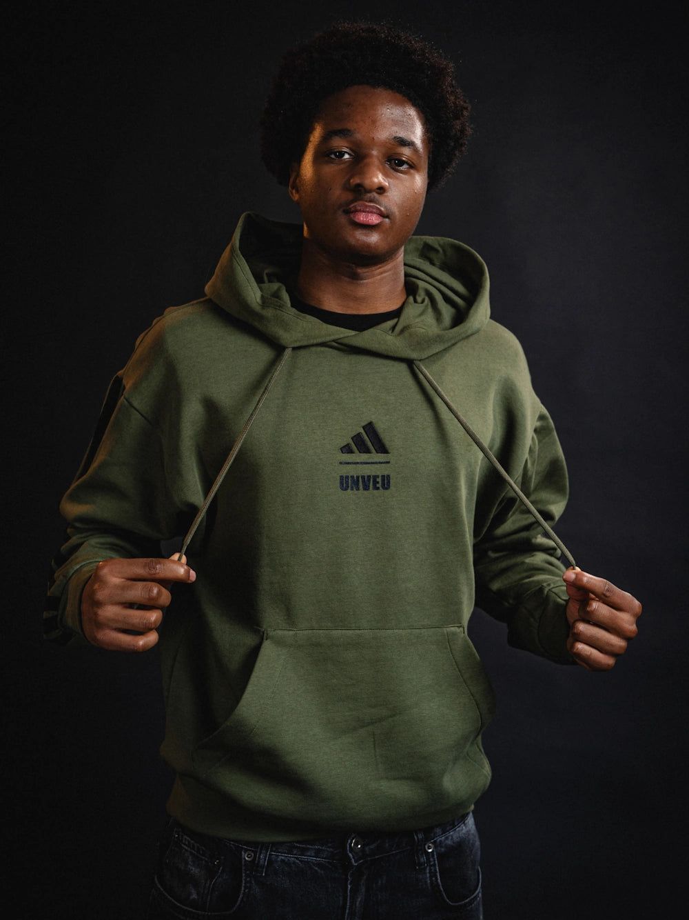Junger Mann trägt einen olivgrünen Hoodie mit Adidas-Logo und der Aufschrift "UNVEIL", vor einem dunklen Hintergrund.