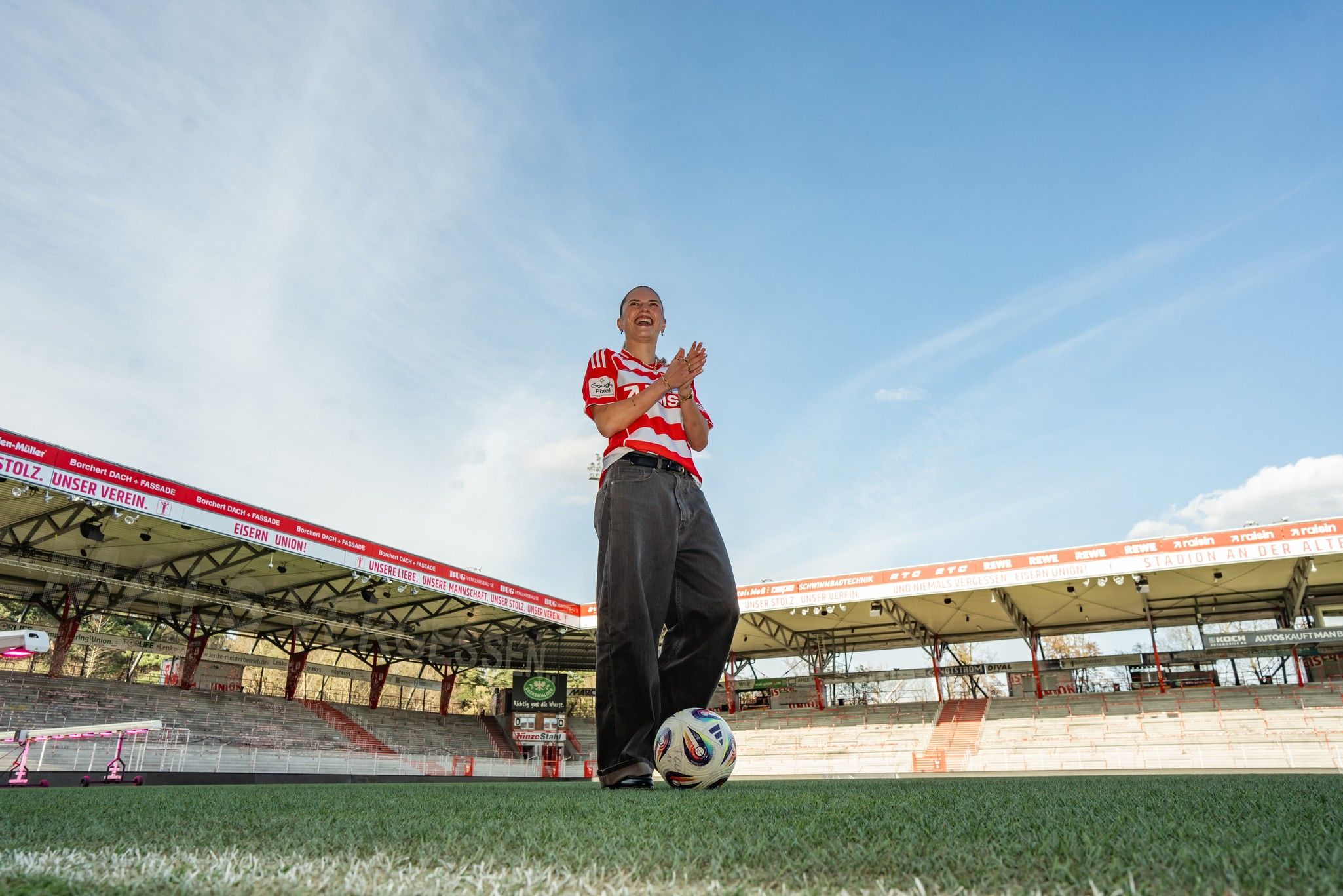 Ein Mann in einem roten Fußballtrikot steht lächelnd auf einem Fußballfeld und applaudiert, mit einem Ball zu seinen Füßen und einem Stadion im Hintergrund.