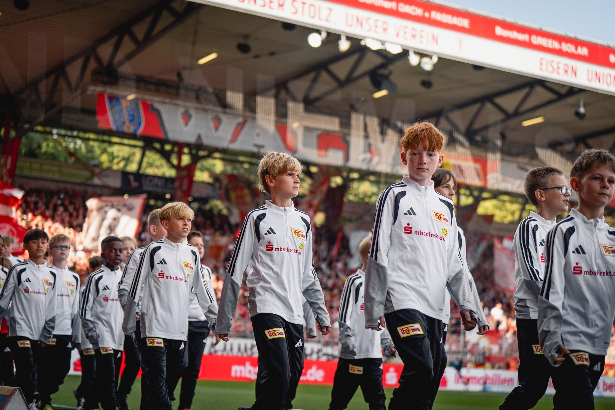Eine Gruppe von Kindern in weißen Sportjacken läuft auf ein Stadionfeld. Hintergrund: Fans und Stadionatmosphäre.