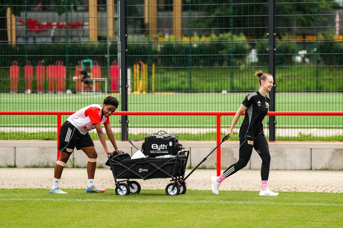 Zwei Personen ziehen einen Wagen auf einem Sportplatz, während im Hintergrund Spielfelder und Übungsgeräte sichtbar sind.