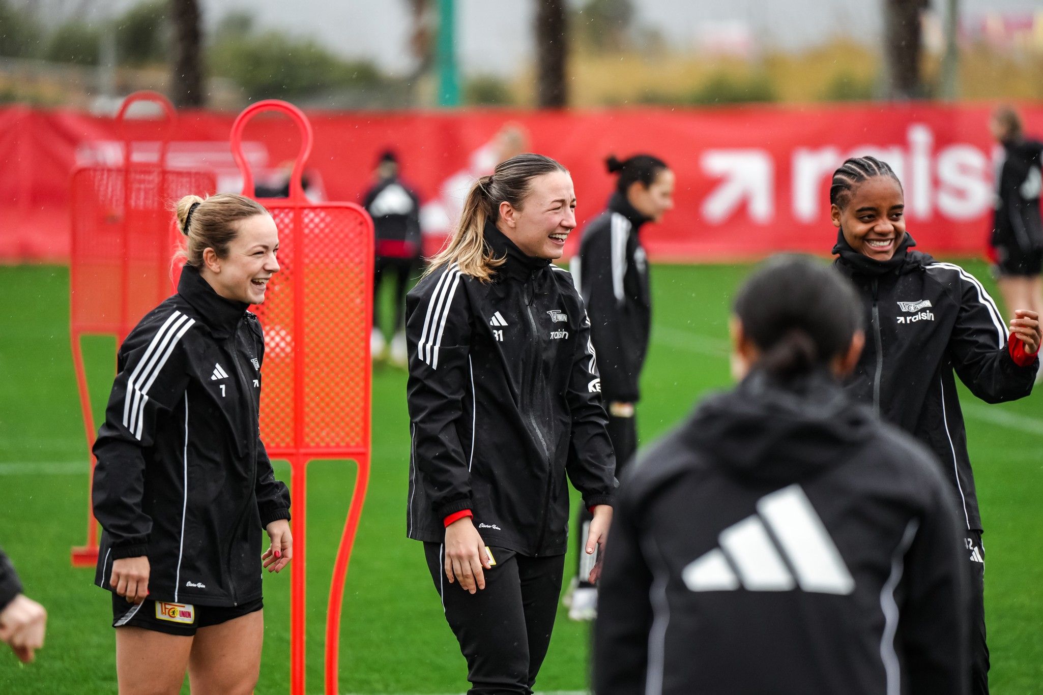 Drei Fußballspielerinnen in Trainingsanzügen lachen und unterhalten sich auf einem Trainingsfeld, während es leicht regnet.