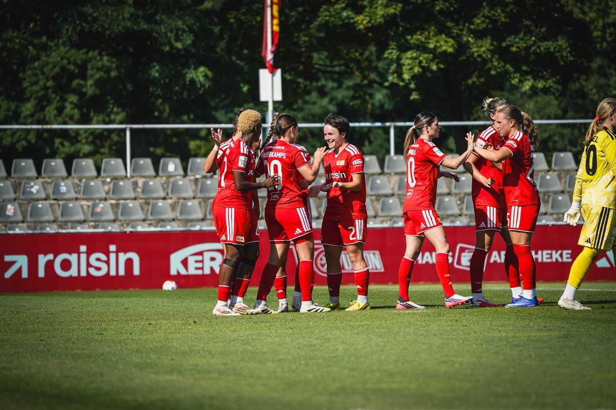 Sechs weibliche Fußballspieler in roten Trikots feiern einen Torerfolg auf dem Spielfeld, im Hintergrund stehlen leere Sitzreihen.