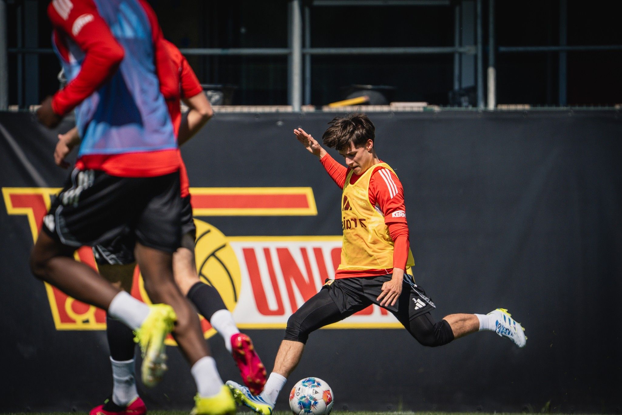 Ein Fußballspieler in gelbem Trainingsshirt führt einen Schuss auf den Ball aus, umgeben von weiteren Spielern im Training.