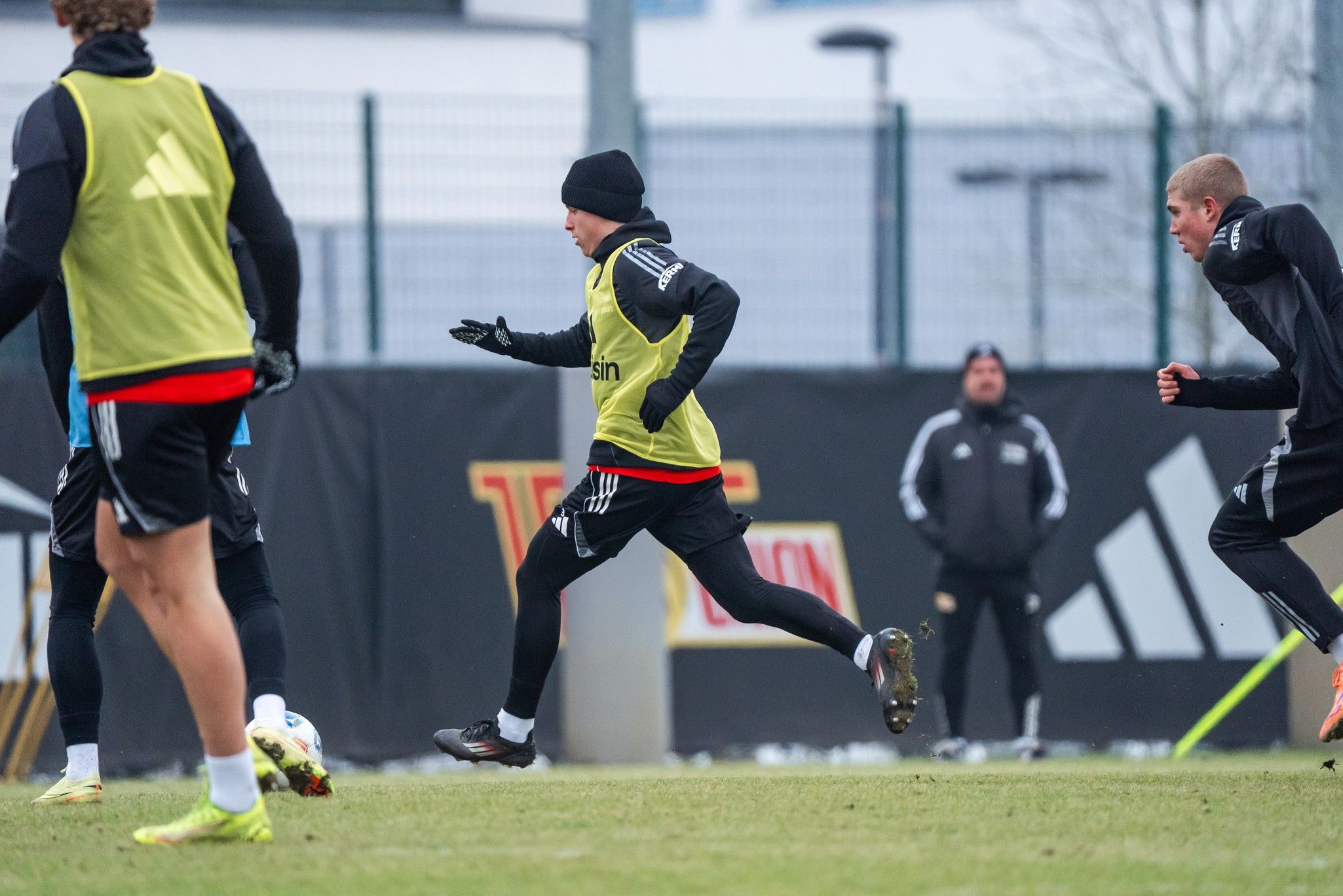 Ein Fußballspieler in einem gelben Trainingsoberteil sprintet auf einem Trainingsplatz, während andere Spieler und ein Trainer zuschauen.