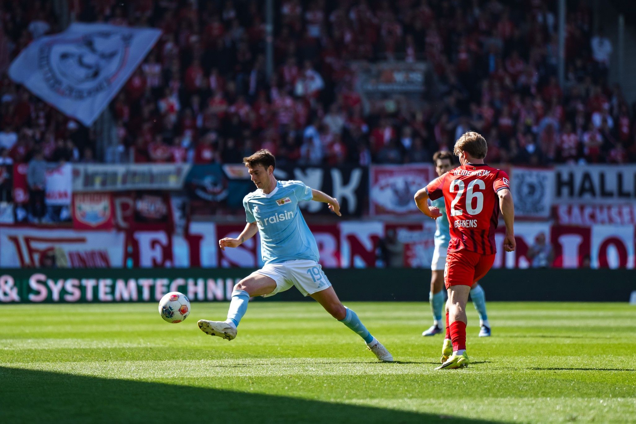 Ein Fußballspieler tritt einen Ball, während ein gegnerischer Spieler ihm gegenübersteht. Im Hintergrund sind Zuschauer und Vereinsfahnen zu sehen.