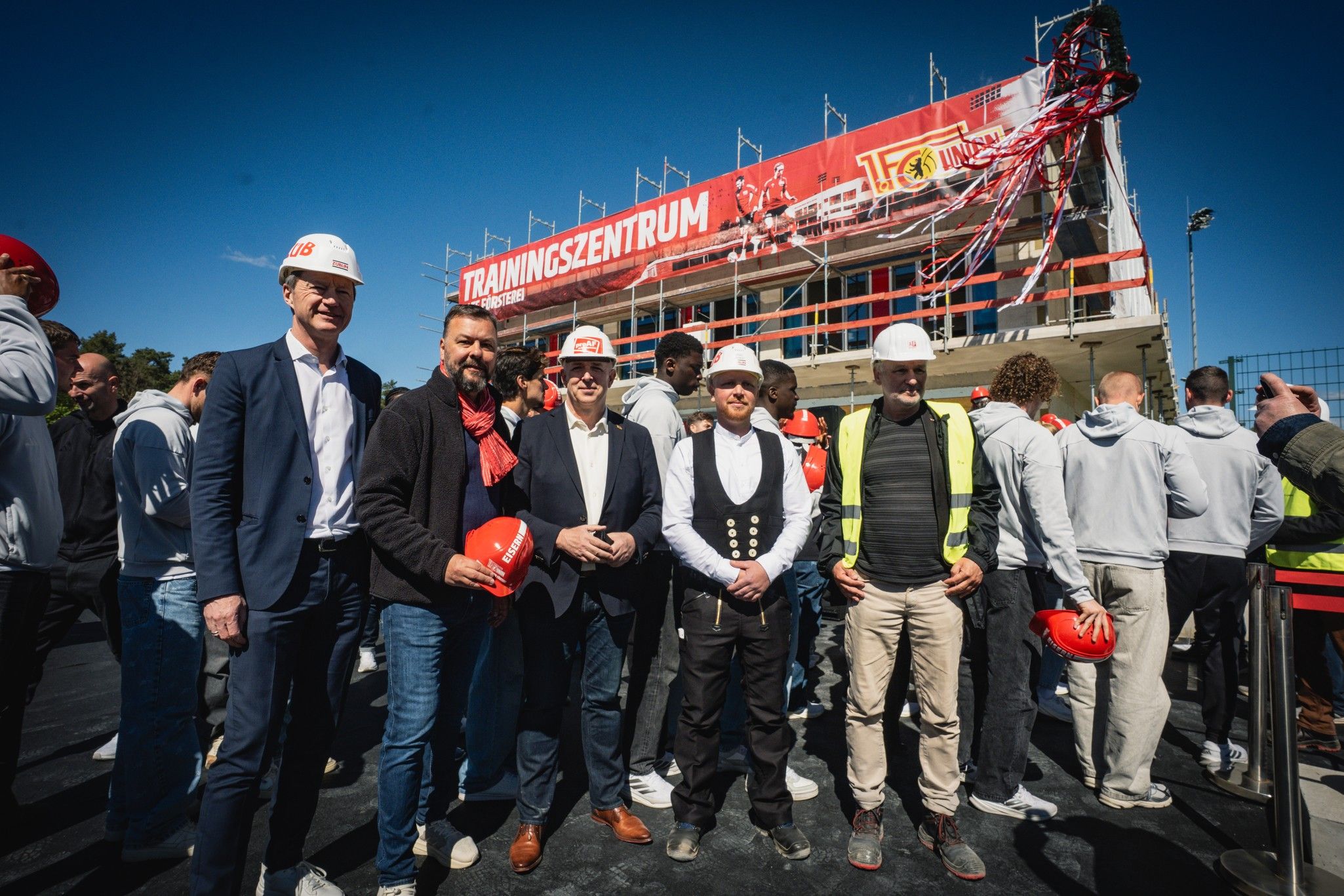 Fünf Männer in Sicherheitshelmen stehen vor einem neu gebauten Trainingszentrum, umgeben von Bauarbeiten und einem blauen Himmel.